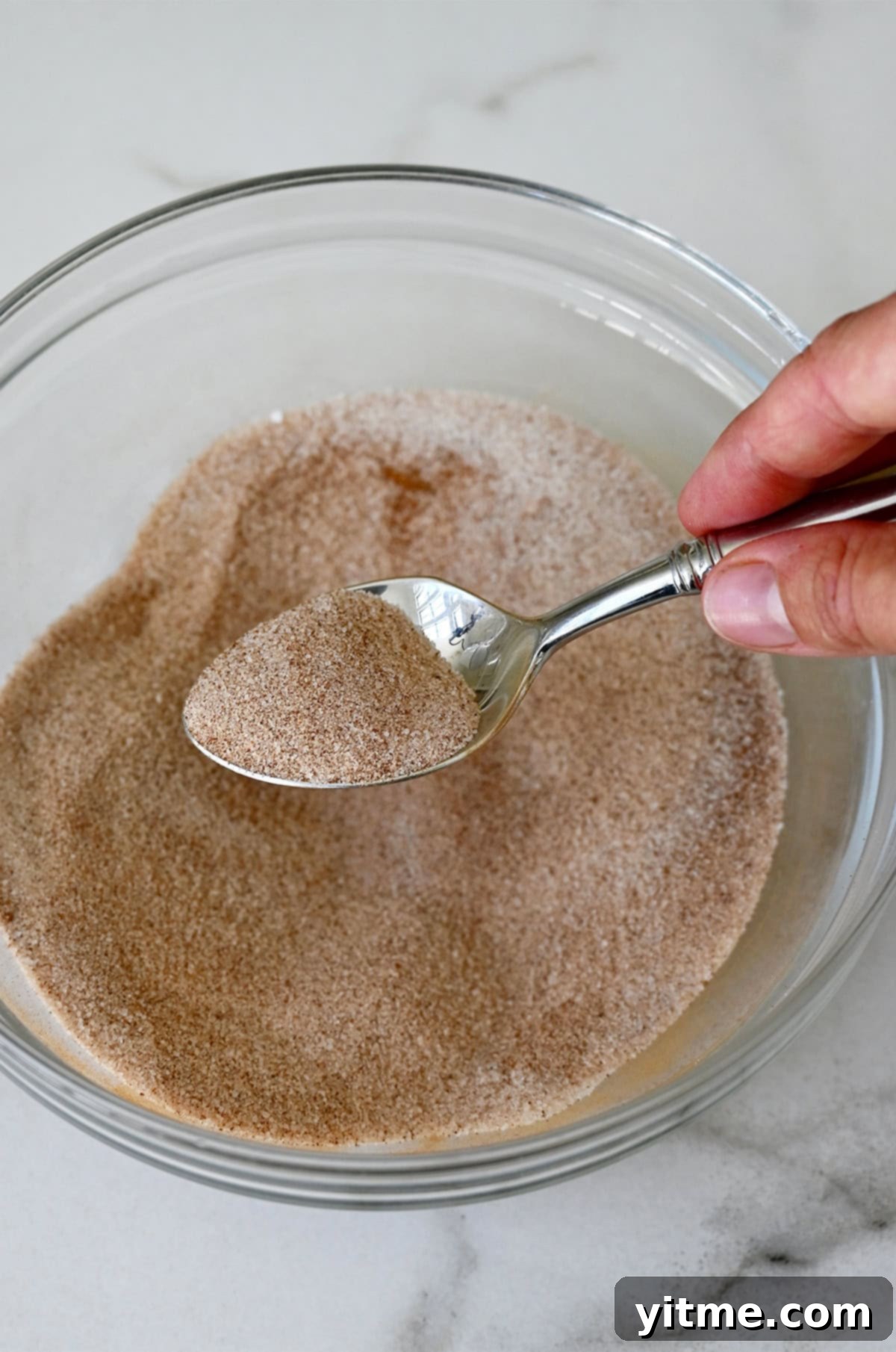 A spoon with cinnamon sugar over a glass bowl with the cinnamon sugar mixture.