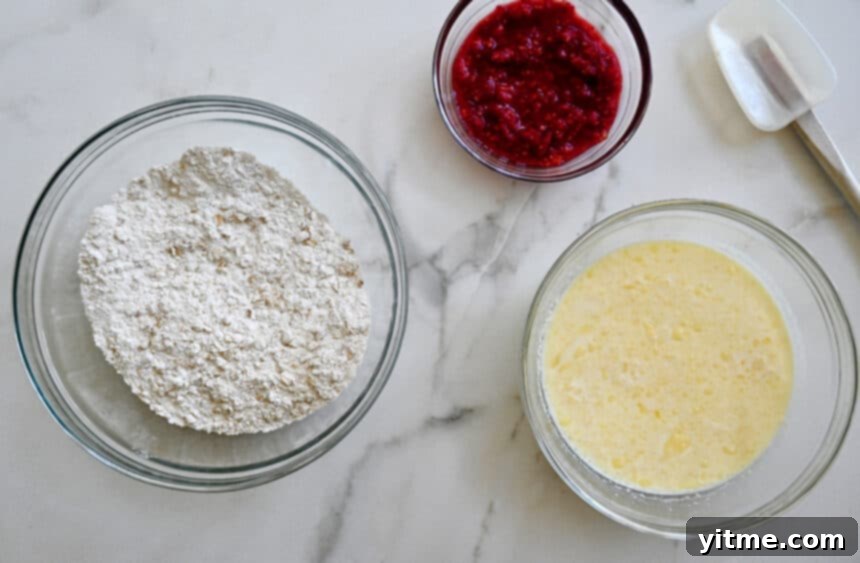 A top-down view of three bowls containing wet and dry ingredients and mashed berries