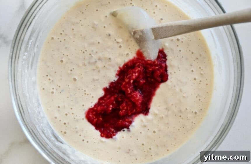Mashed berries being stirred into batter with a spatula