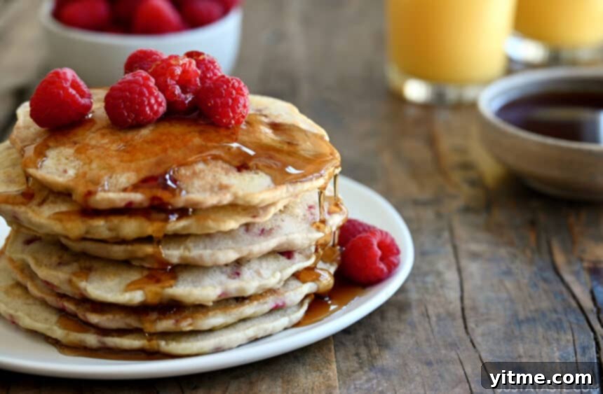 A tall stack of Raspberry Oatmeal Pancakes topped with fresh raspberries and drizzled with maple syrup