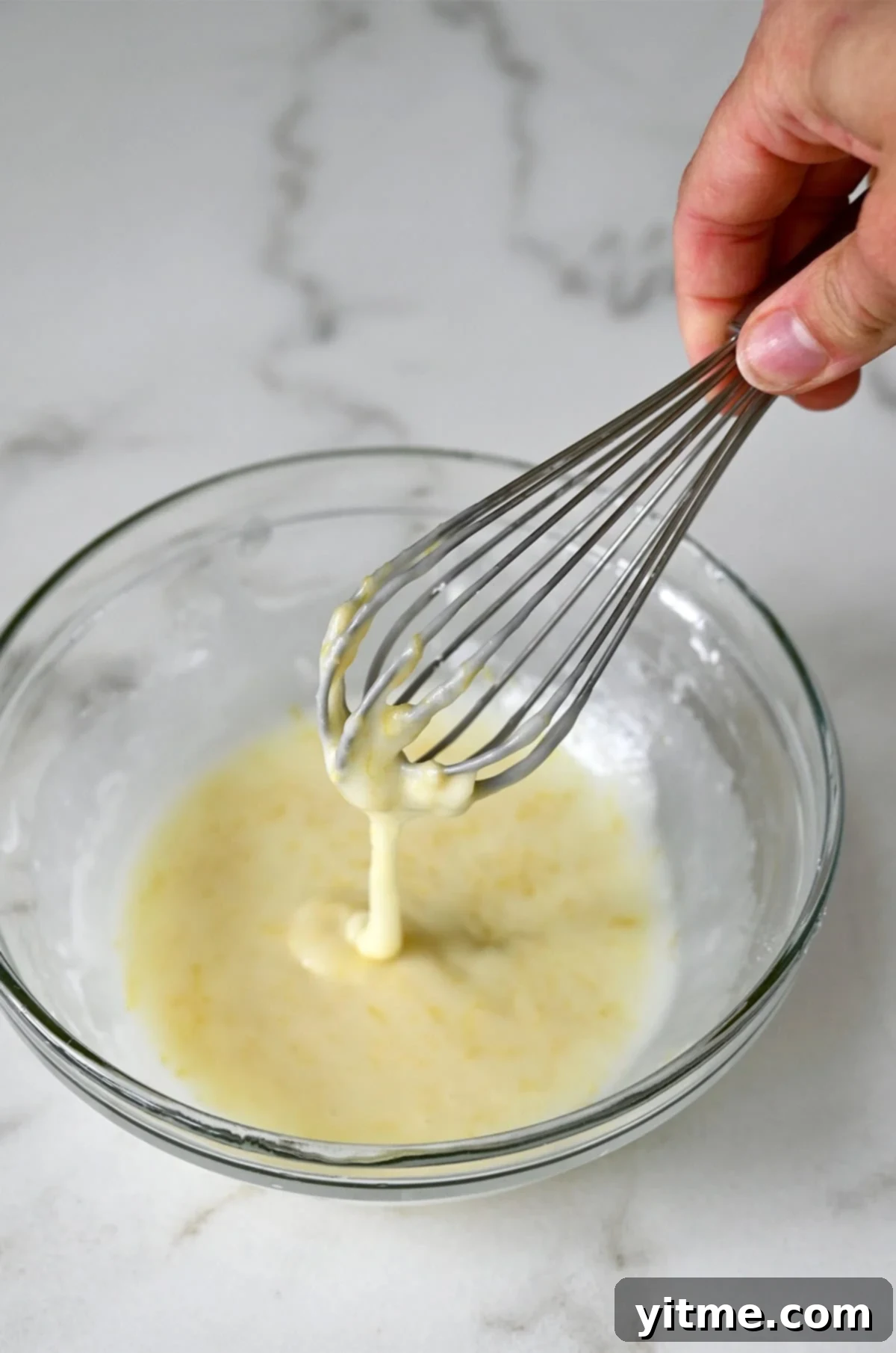 A whisk in a bowl of freshly prepared, smooth lemon glaze, ready to be poured over the bread.