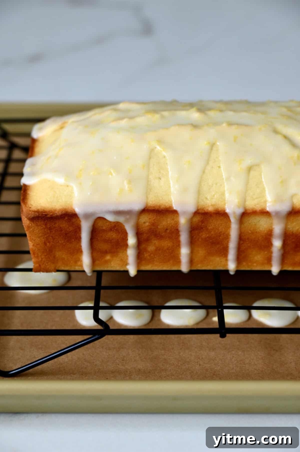 A freshly glazed lemon bread loaf resting on a wire cooling rack, placed over a parchment paper-lined baking sheet to catch excess glaze.