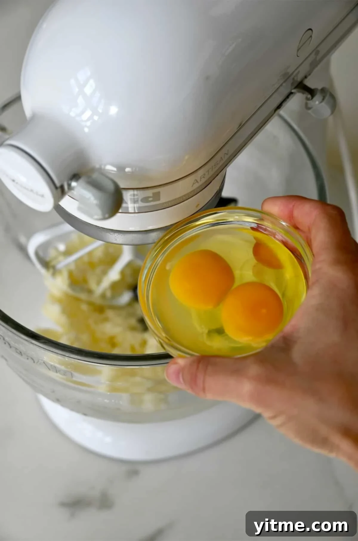 Two eggs being added one by one to the creamed butter and sugar mixture in a stand mixer bowl.