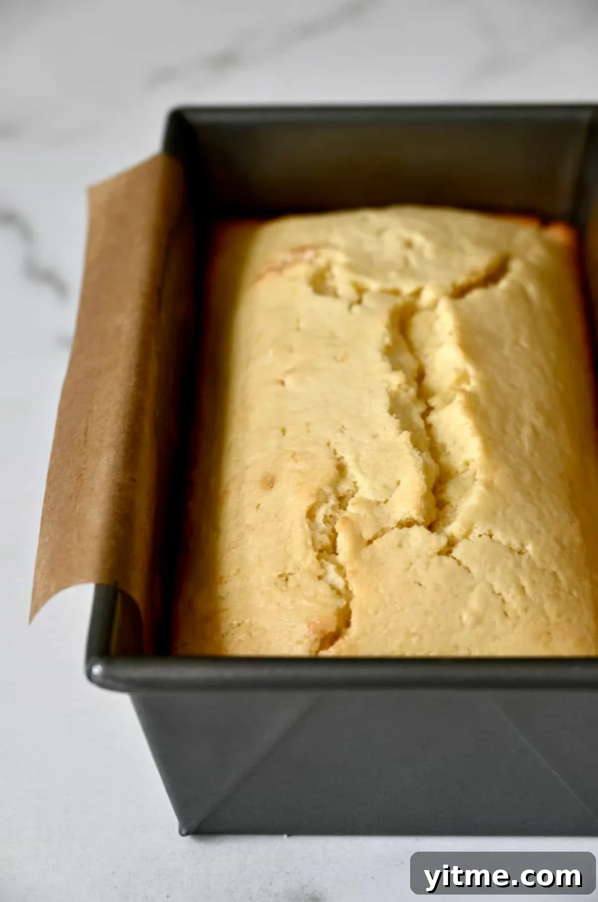A freshly baked golden-brown lemon loaf in a bread pan, cooled slightly on parchment paper.