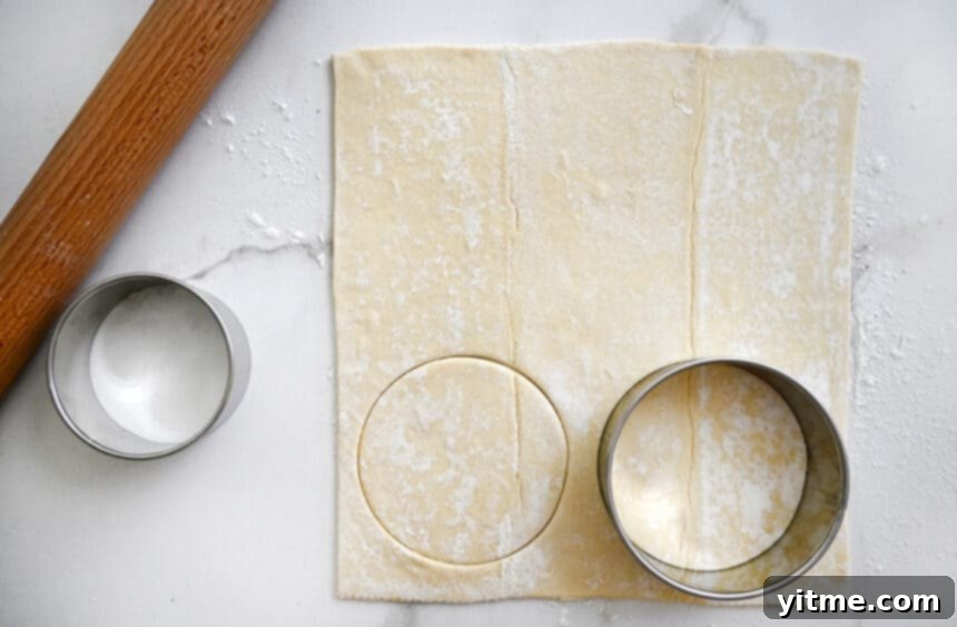 A circular cookie cutter positioned on a sheet of thawed puff pastry, next to a wooden rolling pin, ready for shaping delicious breakfast pastries.