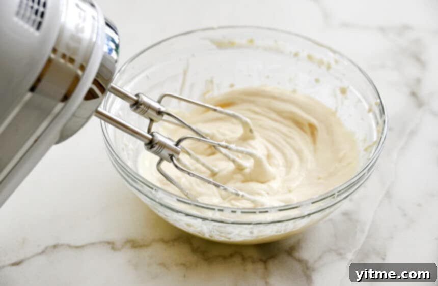 A handheld electric mixer rests against a clear glass bowl, showcasing a perfectly whipped and creamy cream cheese filling, ready to be spread onto breakfast pastries.