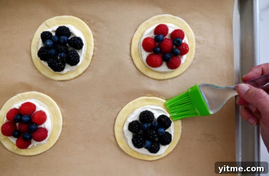A skilled hand uses a pastry brush to apply a shimmering egg wash to the edges of unbaked cream cheese pastries, arranged neatly on a parchment paper-lined baking sheet, enhancing their golden potential.