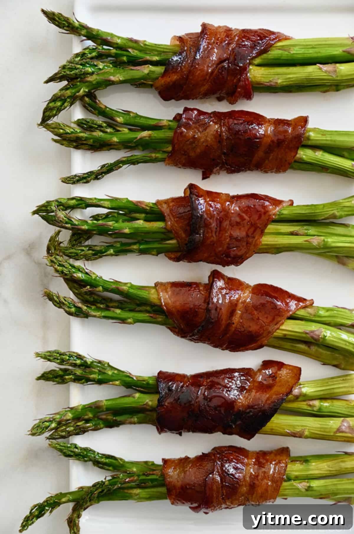 Six cooked bacon-wrapped asparagus bundles on a white platter.