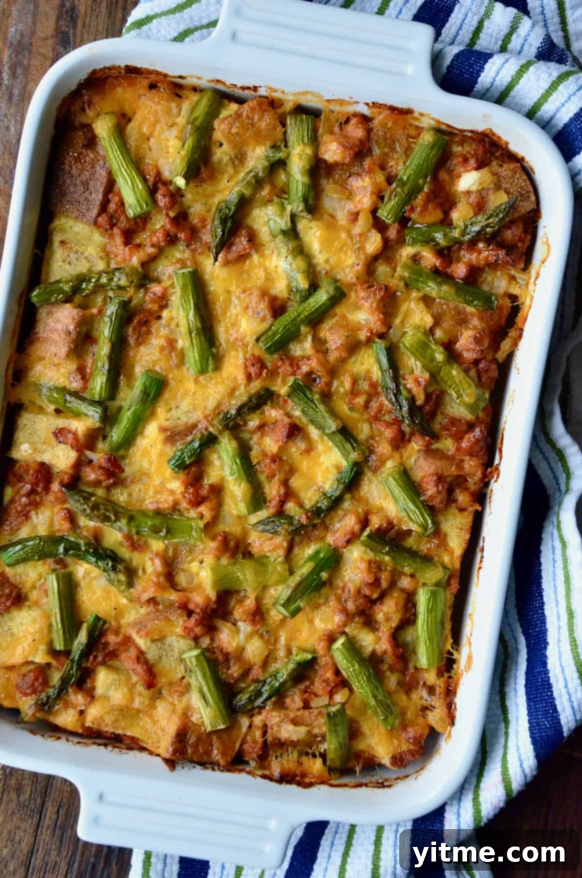 A casserole dish holding a baked breakfast strata with sausage and asparagus. The dish sits on a kitchen towel on a wooden table.