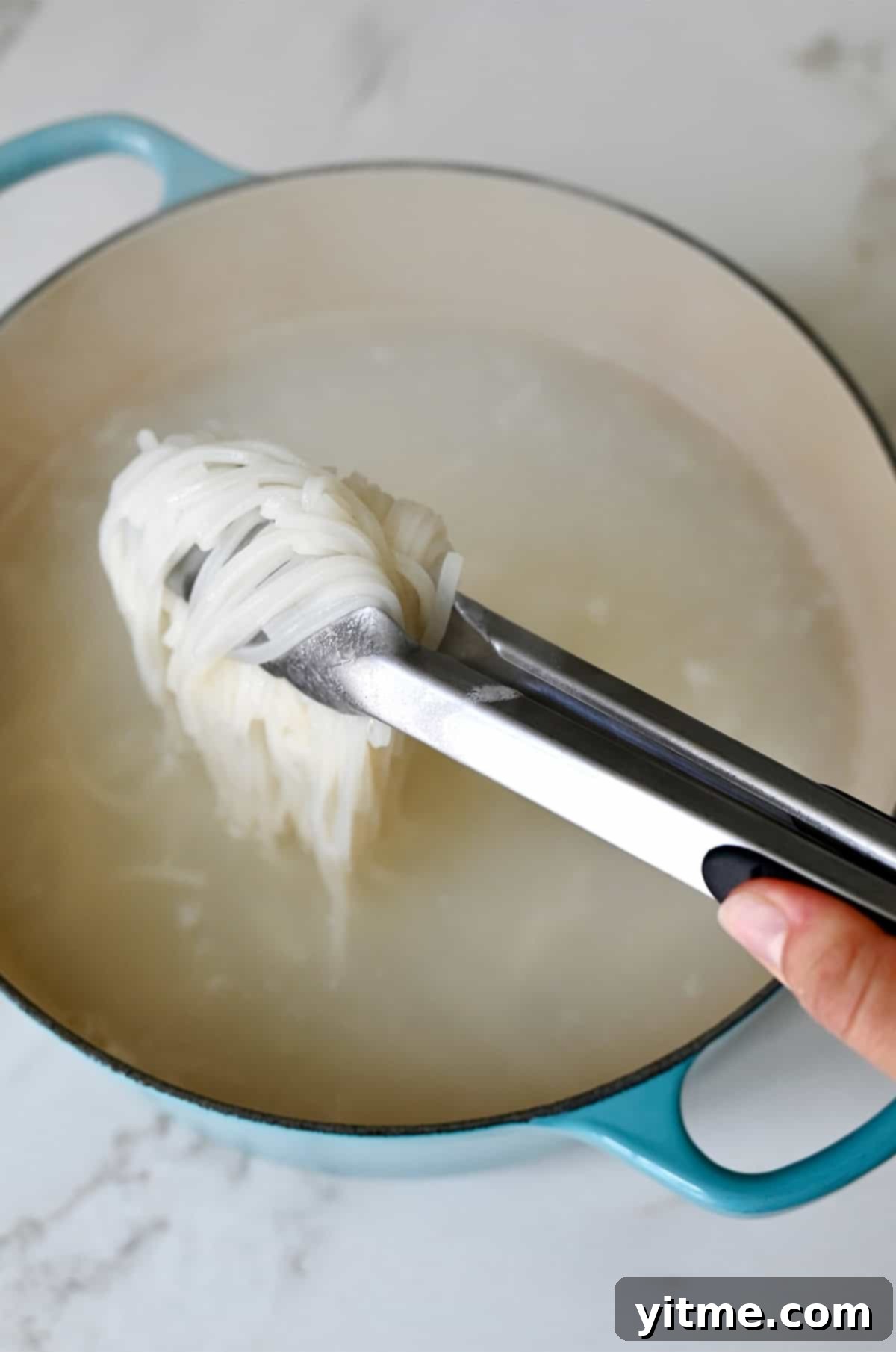 A hand holding tongs pulls cooked rice noodles out of water in a large stockpot.