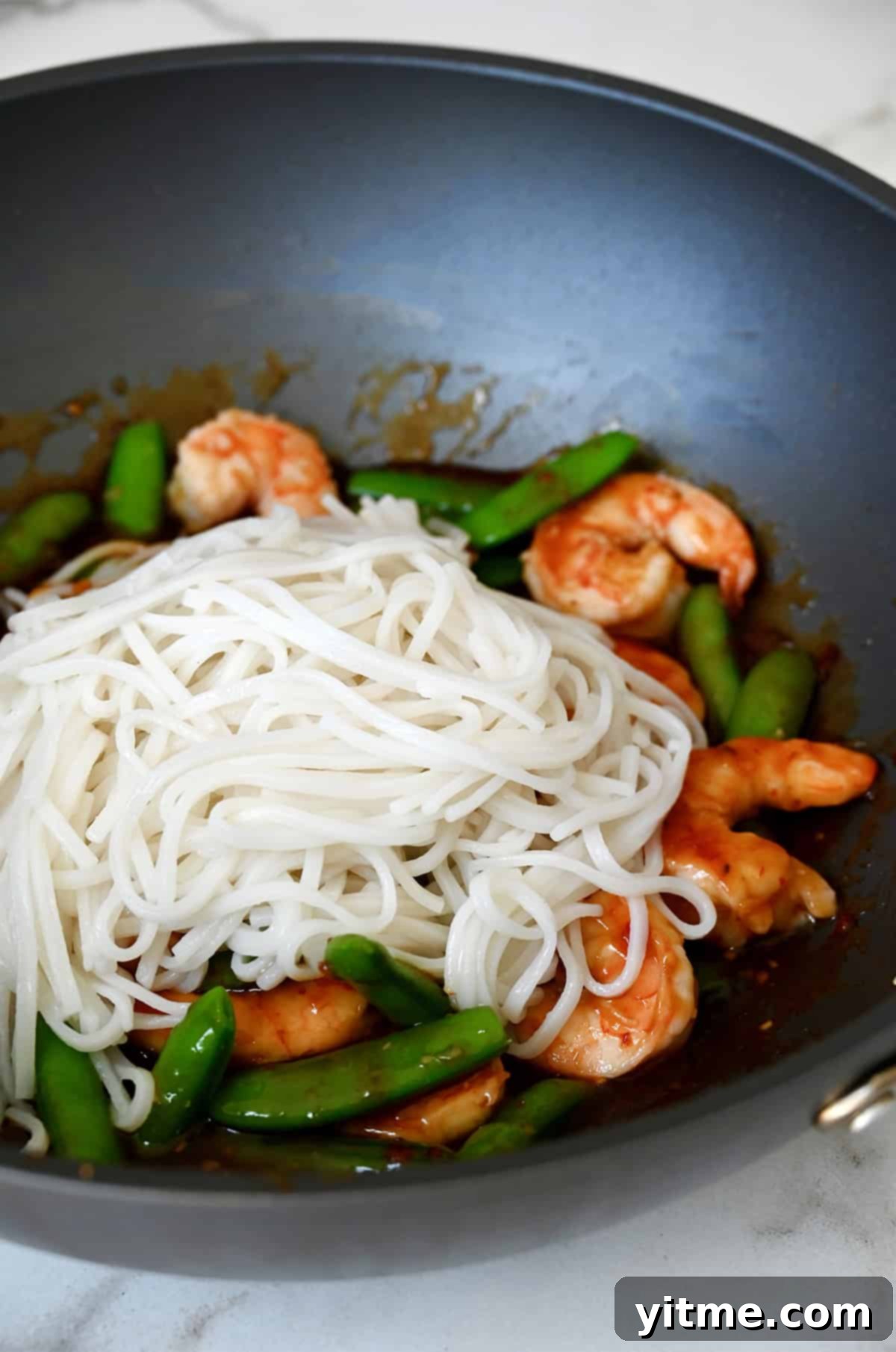 Cooked rice noodles in a wok with sugar snap peas and sweet and sour shrimp.
