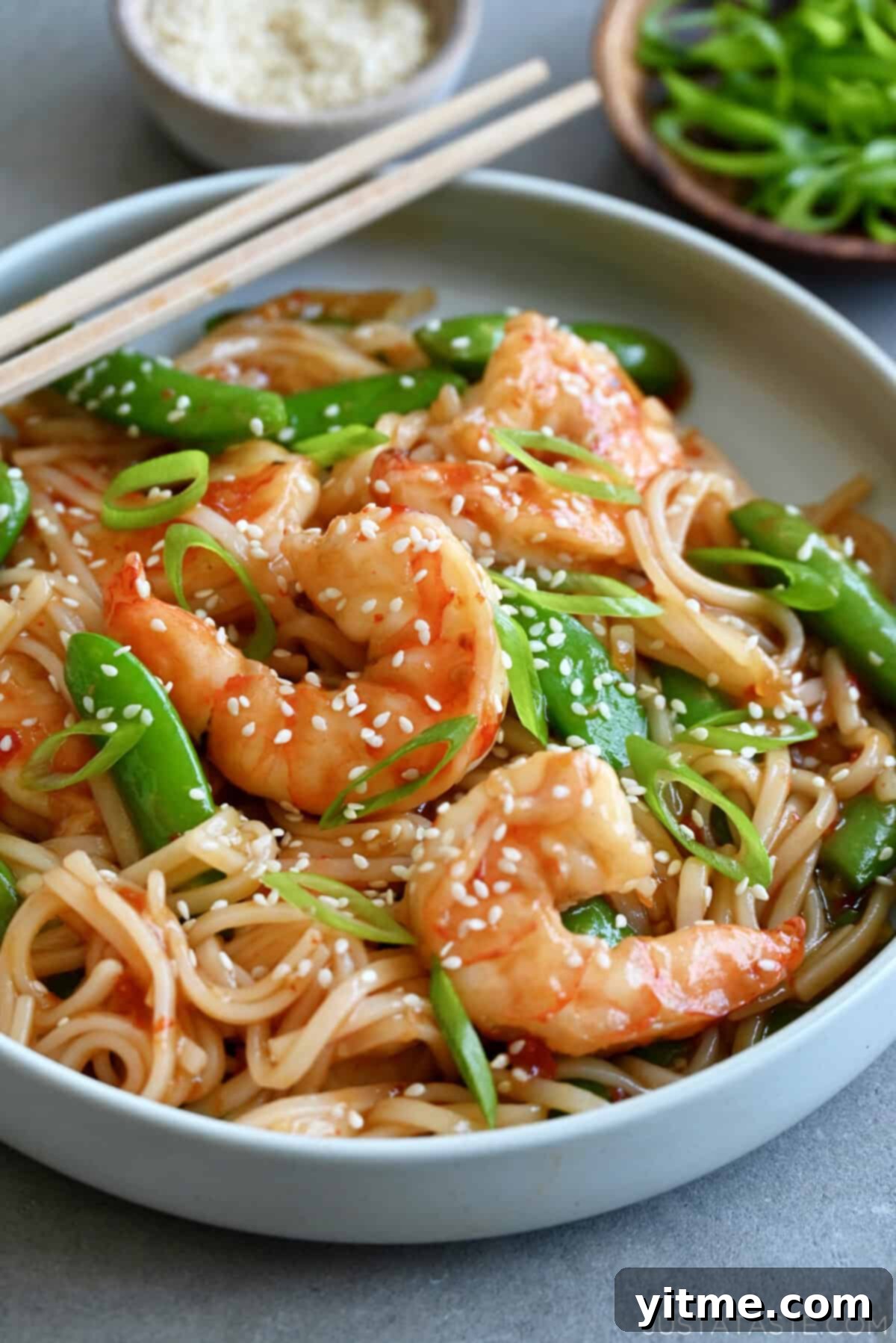 Sweet and sour shrimp stir-fry with rice noodles garnished with sesame seeds in a shallow bowl with chopsticks.