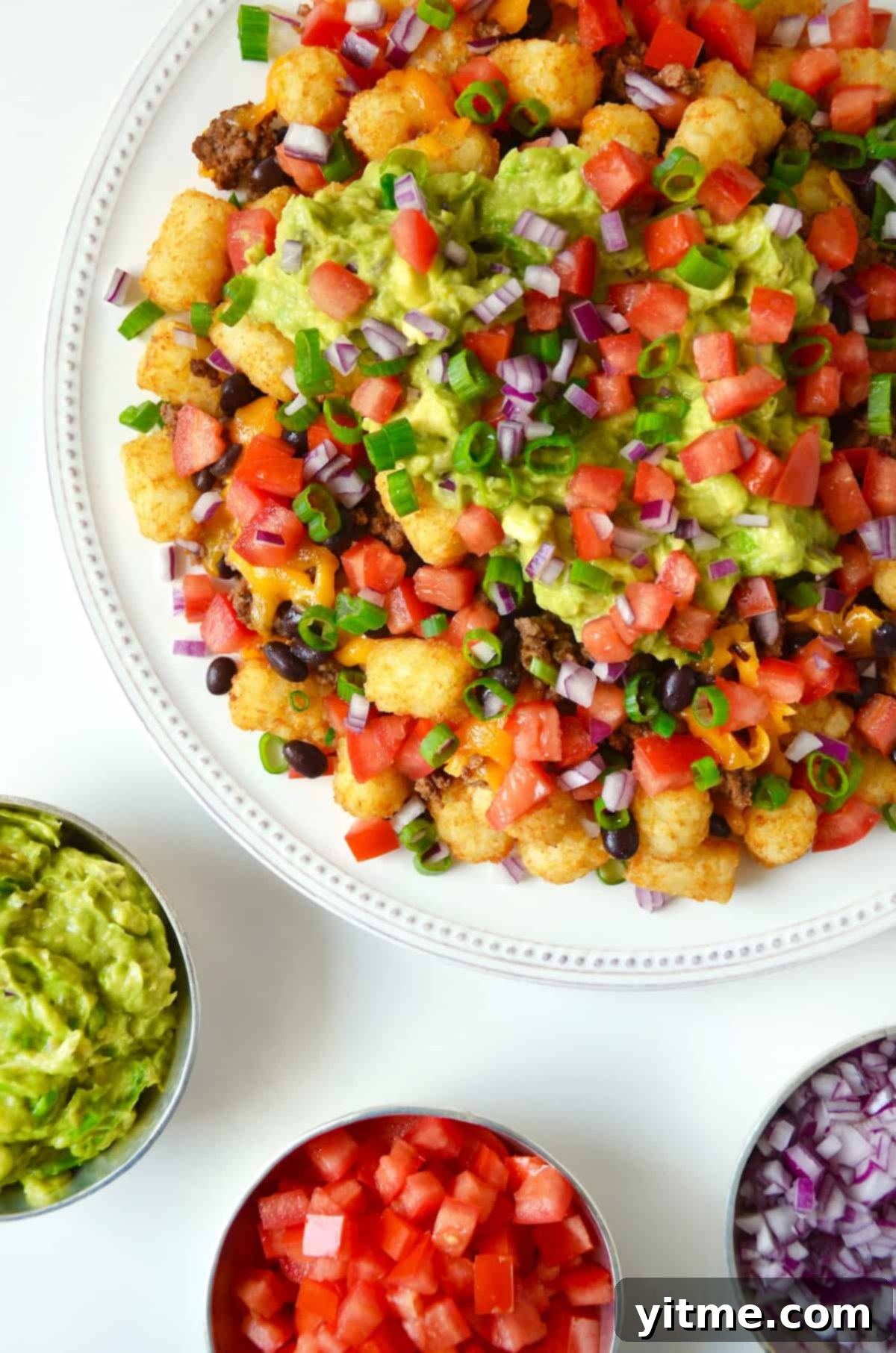 A white serving platter with loaded totchos (tater tot nachos) topped with diced tomatoes, scallions, black beans and diced avocado.