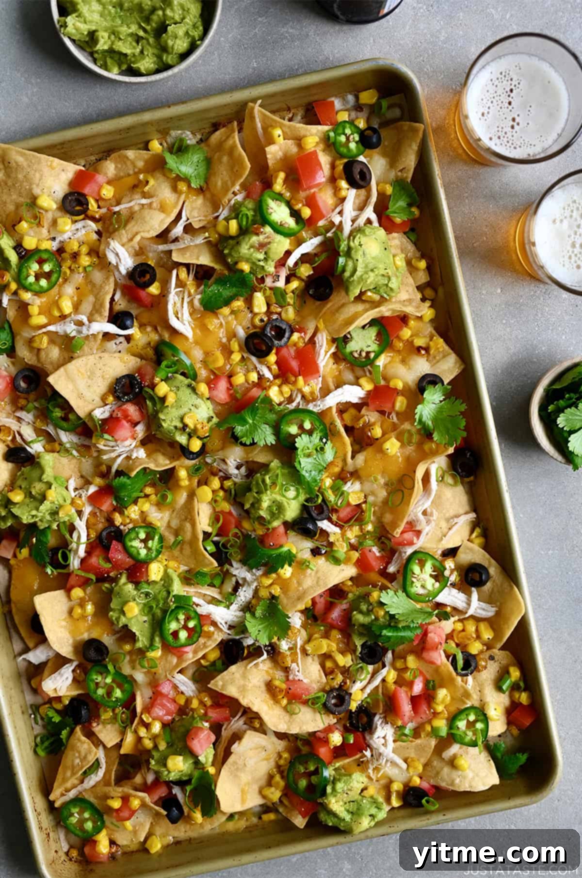 Sheet pan nachos loaded with sliced jalapeños, diced tomatoes, black olives and guacamole next to two glasses filled with light beer.