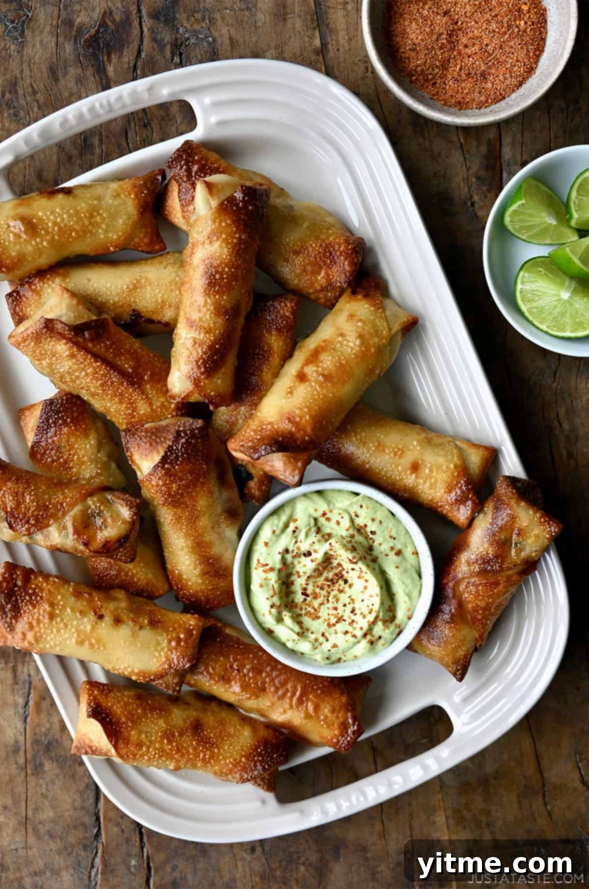 A bird's-eye view of crispy baked egg rolls accompanied by small bowls of creamy avocado dip, zesty Tajin seasoning, and a refreshing lime, creating a flavorful and colorful appetizer spread.