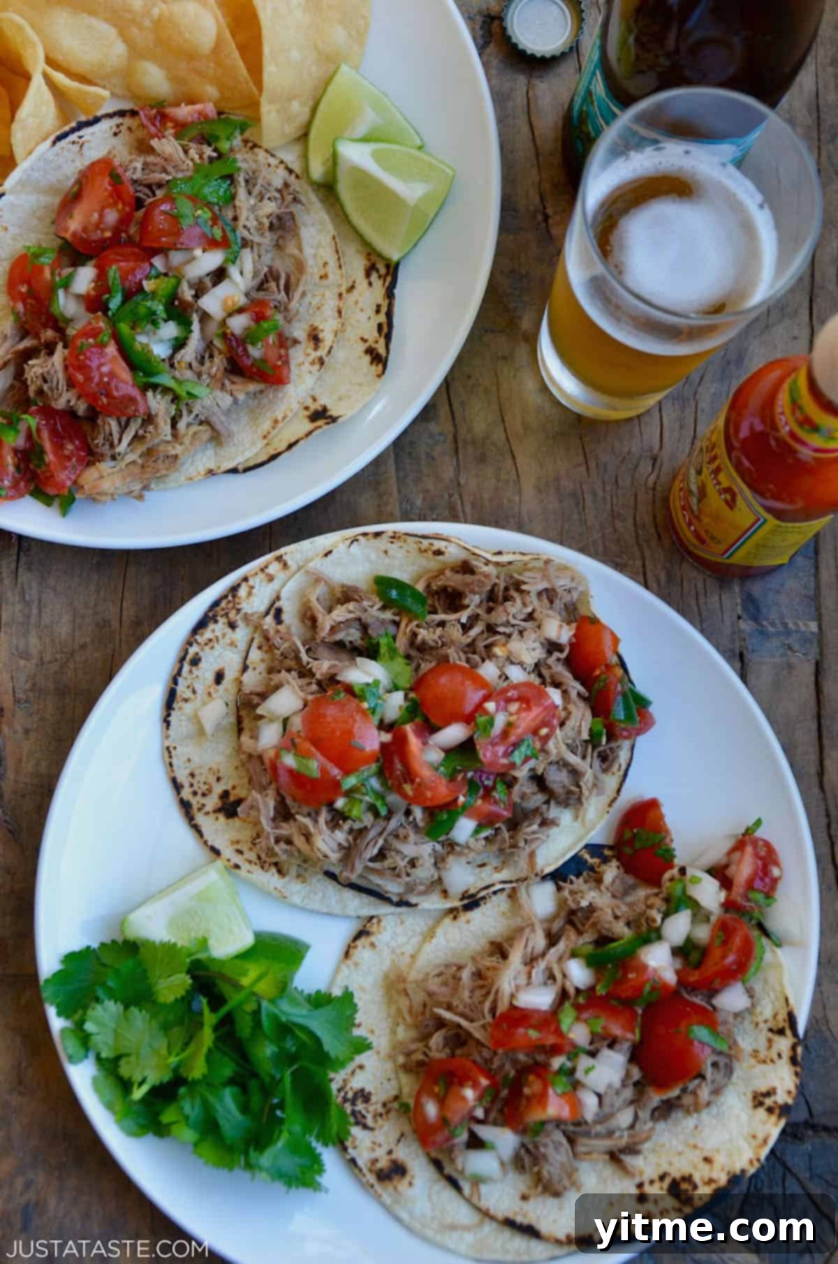 Two plates of tantalizing carnitas tacos topped with vibrant cherry tomato pico de gallo, accompanied by fresh cilantro and zesty lime wedges, alongside a bottle of fiery hot sauce and a refreshing glass of beer.