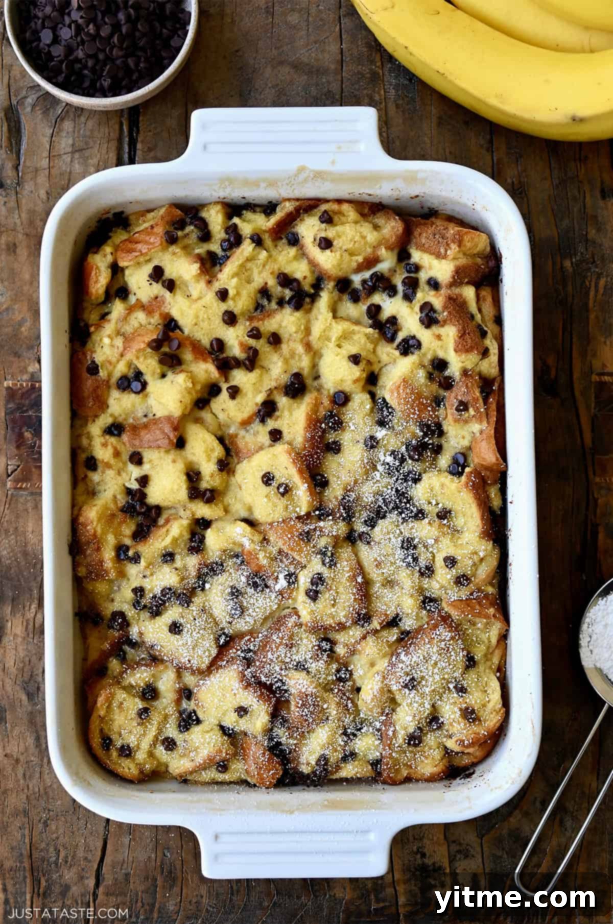Golden brown banana bread pudding with melted chocolate chips, lightly dusted with powdered sugar on top.