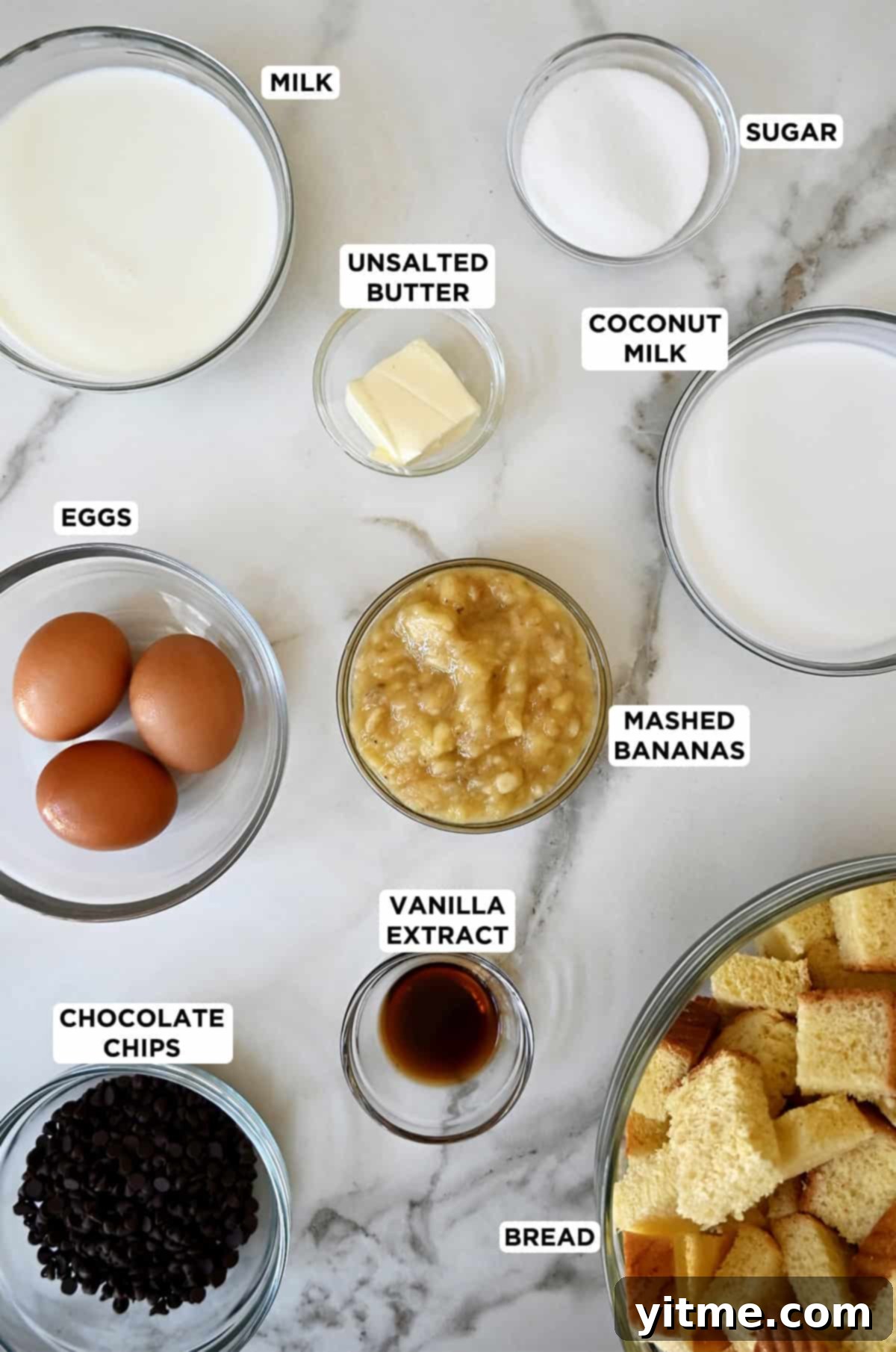 Arrangement of glass bowls displaying all ingredients for banana bread pudding: coconut milk, eggs, chocolate chips, mashed bananas, vanilla extract, milk, cubed brioche bread, and butter.