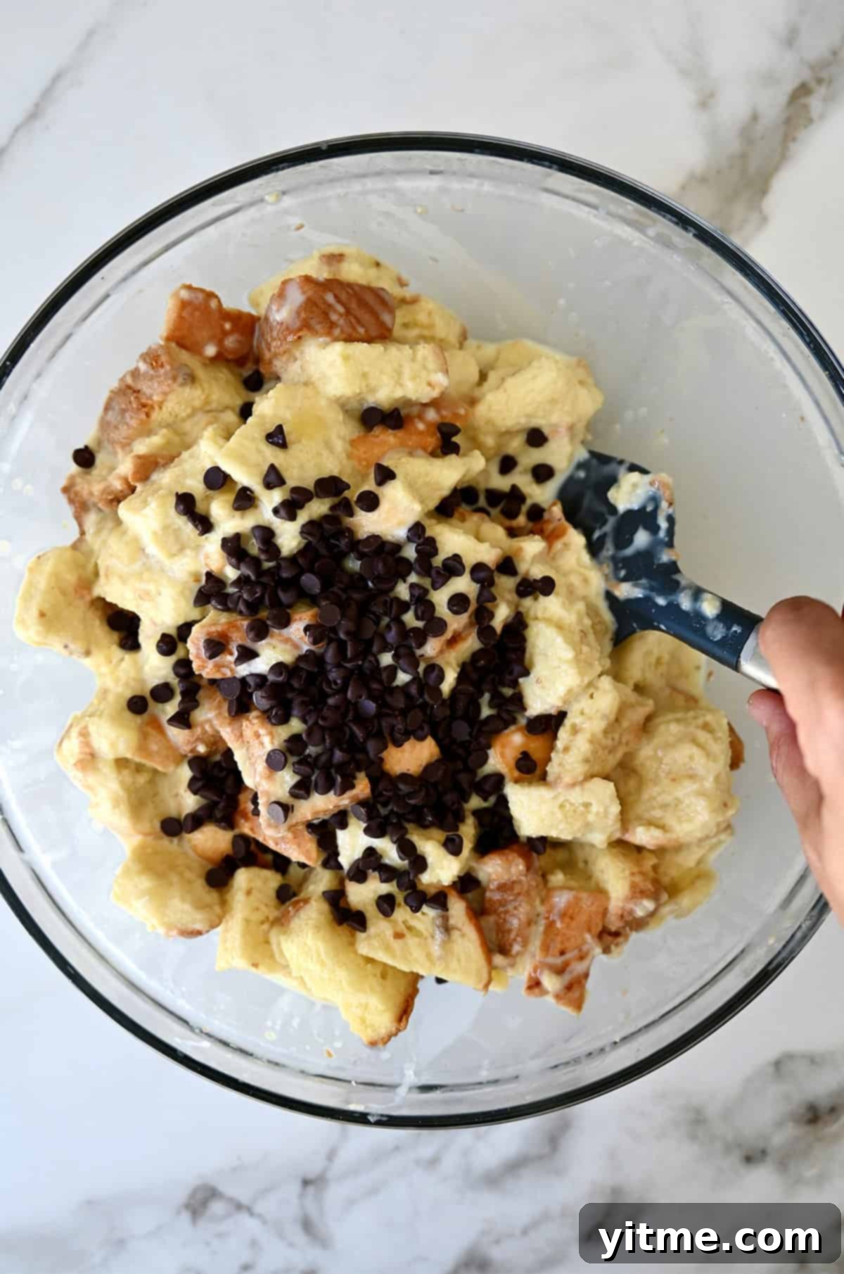Mini chocolate chips atop custard-soaked cubes of bread in a glass bowl, ready to be folded in.
