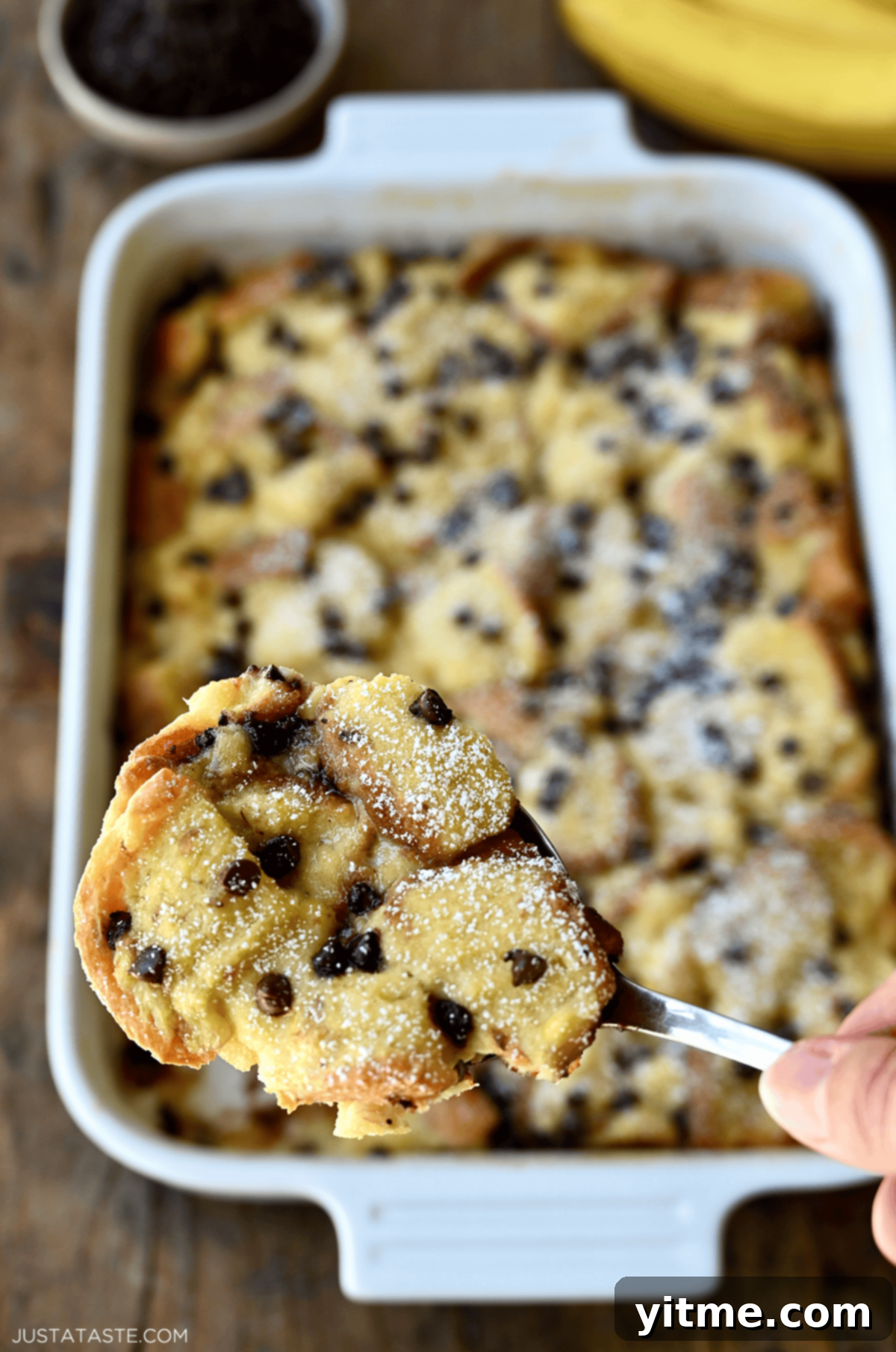 A hand holds a spoon with a big scoop of chocolate chip banana bread pudding on it, showing gooey chocolate and soft bread.