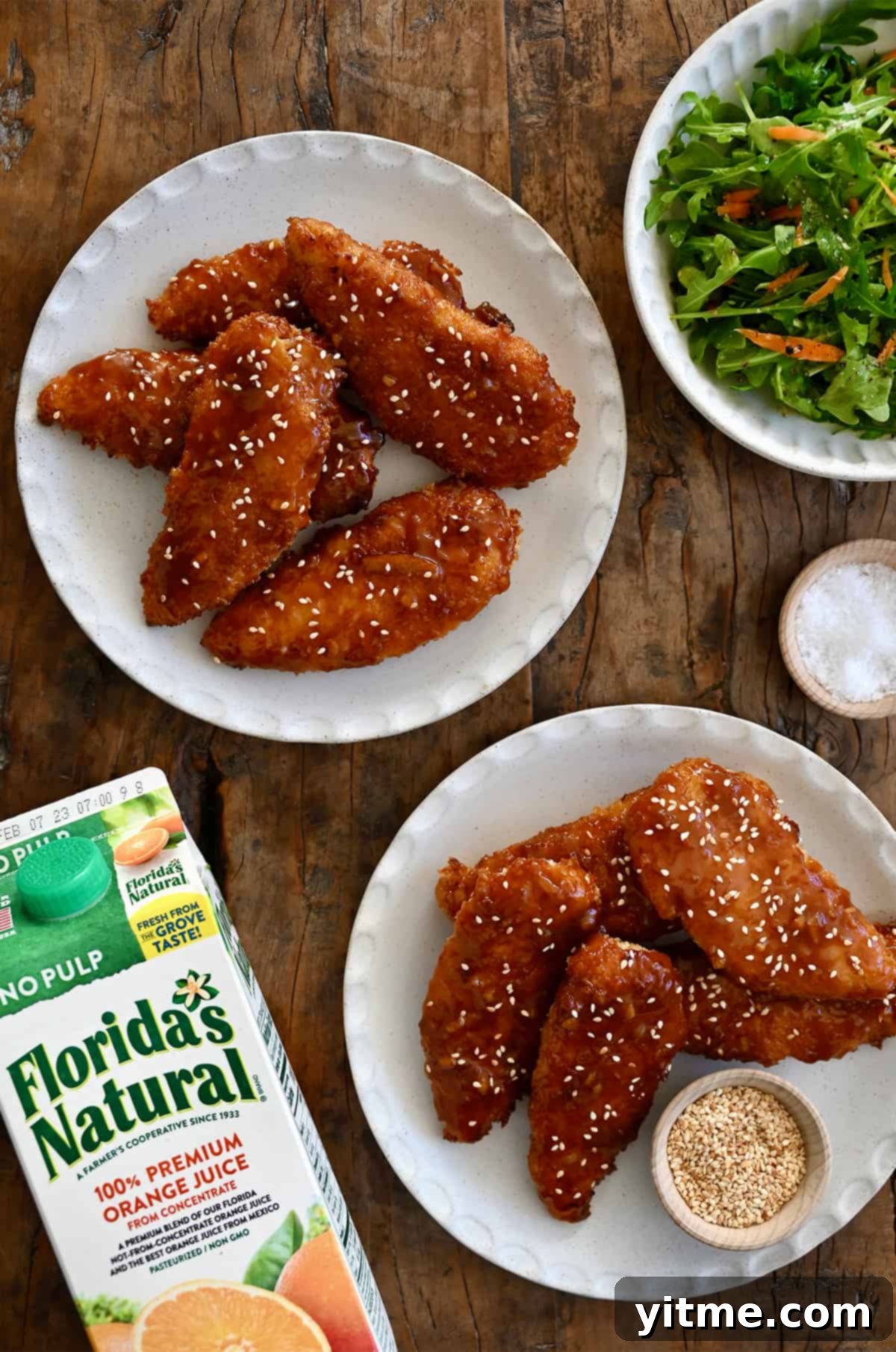 Citrus Glazed Baked Chicken Strips 2 A top-down view of two plates piled high with orange chicken tenders next to a carton of Florida's Natural Orange Juice, garnished with sesame seeds.