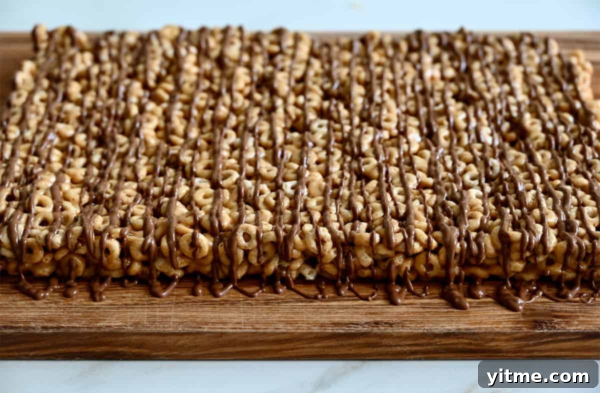 Peanut Butter Cheerios Marshmallow Bars 5 Freshly made marshmallow Cheerios bars, generously drizzled with melted chocolate, resting on a cutting board, ready to be sliced into perfect squares.