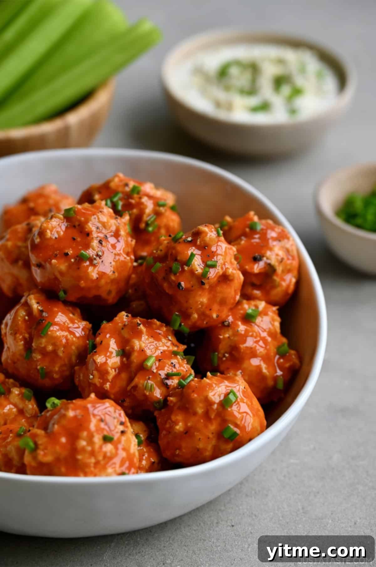 Buffalo chicken meatballs in a bowl garnished with chopped scallions near a bowl containing blue cheese dressing.