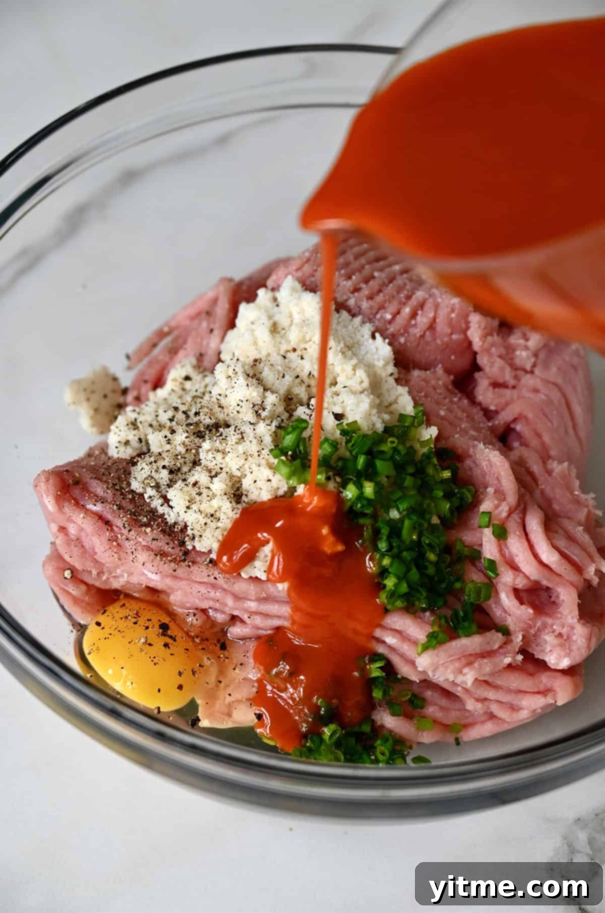 Buffalo sauce being poured over ground chicken, milk-soaked breadcrumbs, chopped chives and an egg in a large bowl.