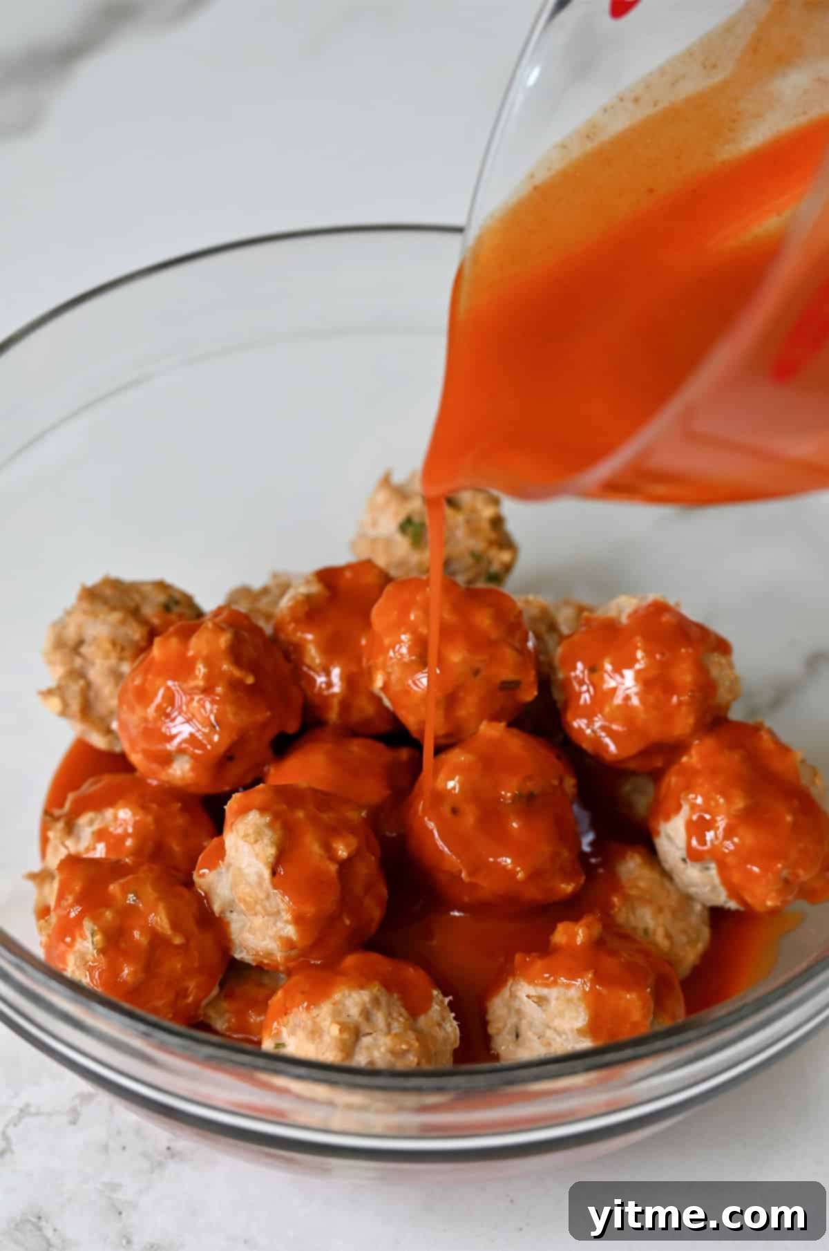 Buffalo sauce being poured over mini chicken meatballs in a bowl.