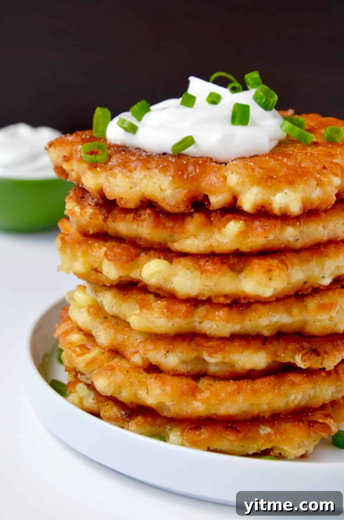 A tall stack of corn fritters topped with a dollop of sour cream and garnished with chopped scallions.