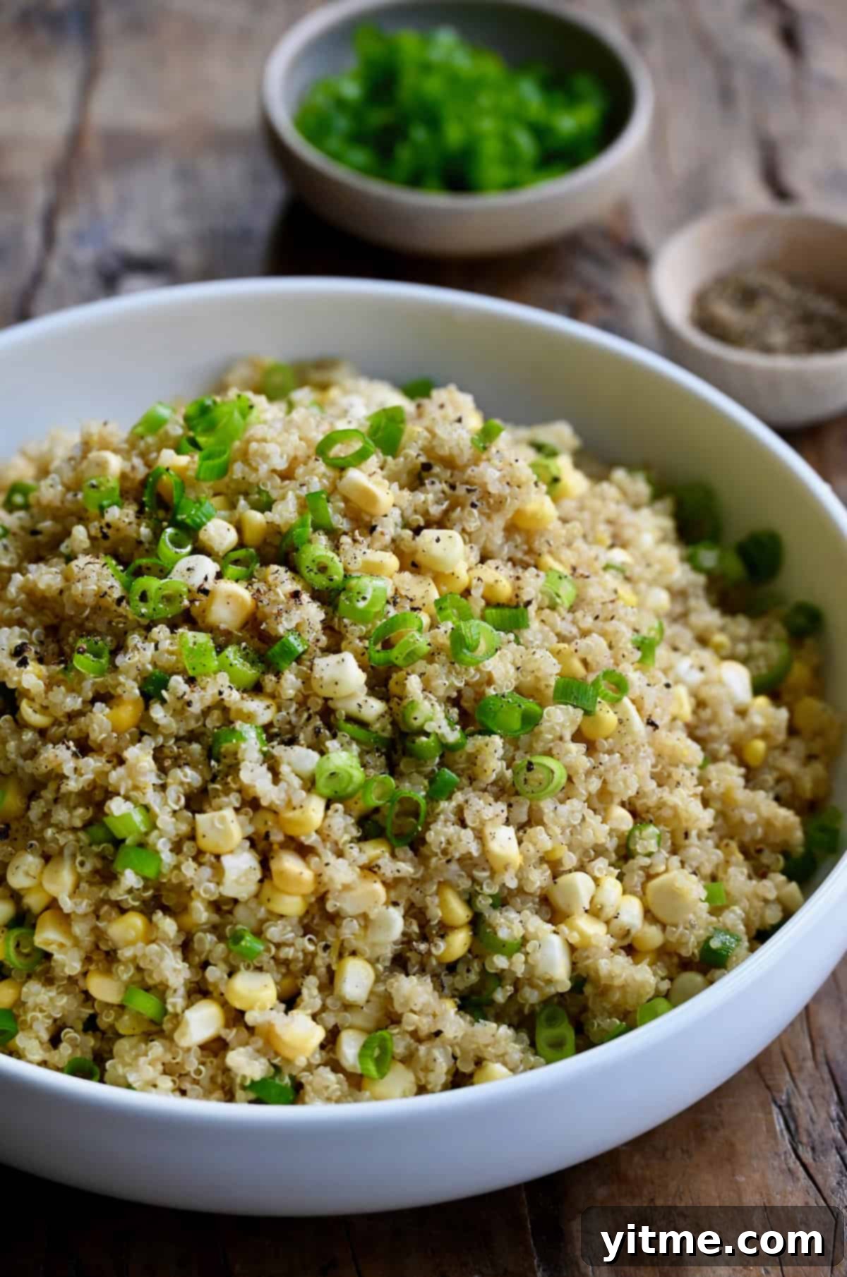 Quinoa with corn and scallions in a large white serving bowl. Small bowls of sliced scallions and ground black pepper are behind the serving bowl.