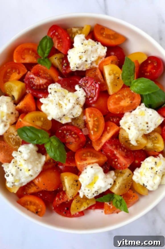 A white bowl containing red, orange and yellow halved tomatoes, burrata and basil
