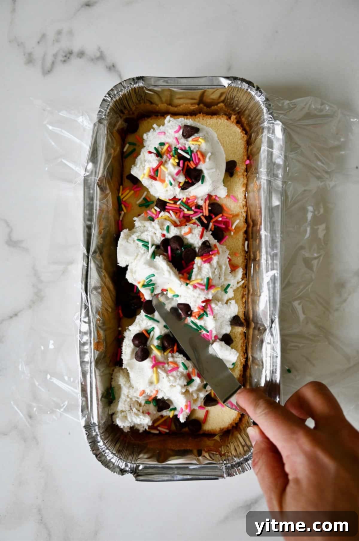 A hand holding an offset spatula spreads ice cream atop pound cake in a aluminum pan lined with plastic wrap.