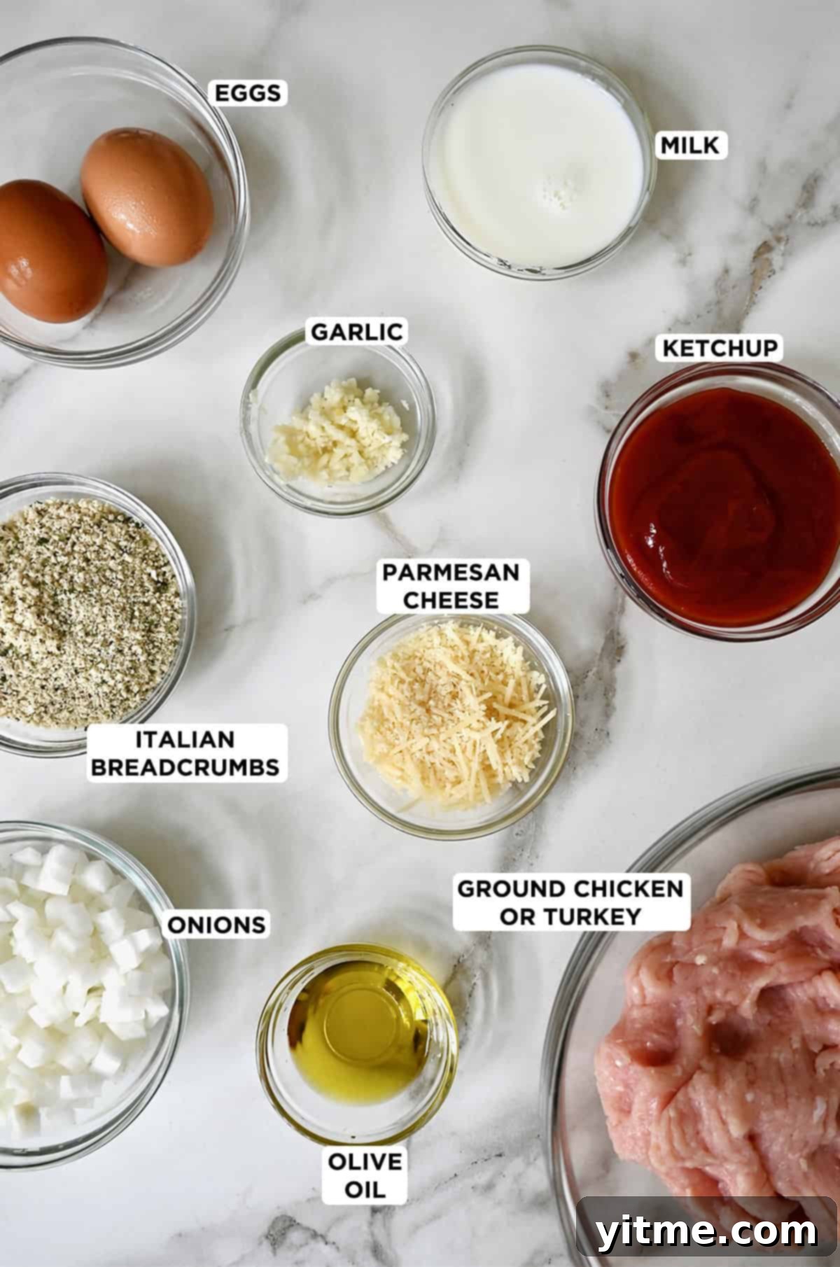 Glass bowls arranged with fresh ingredients for making chicken meatloaf, including ground chicken, Italian breadcrumbs, finely minced white onions and garlic, grated Parmesan cheese, classic ketchup, eggs, olive oil, and milk.