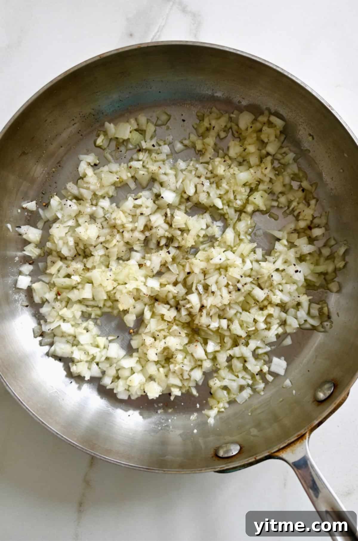 Finely minced white onions being sautéed in a skillet with a drizzle of olive oil until translucent and fragrant.