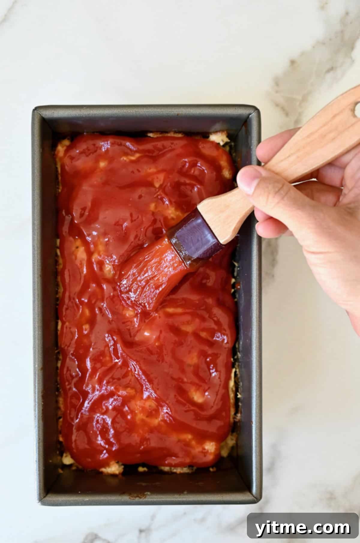 A hand expertly applying a vibrant ketchup glaze over the top of a partially baked chicken meatloaf using a pastry brush, ensuring even coverage.