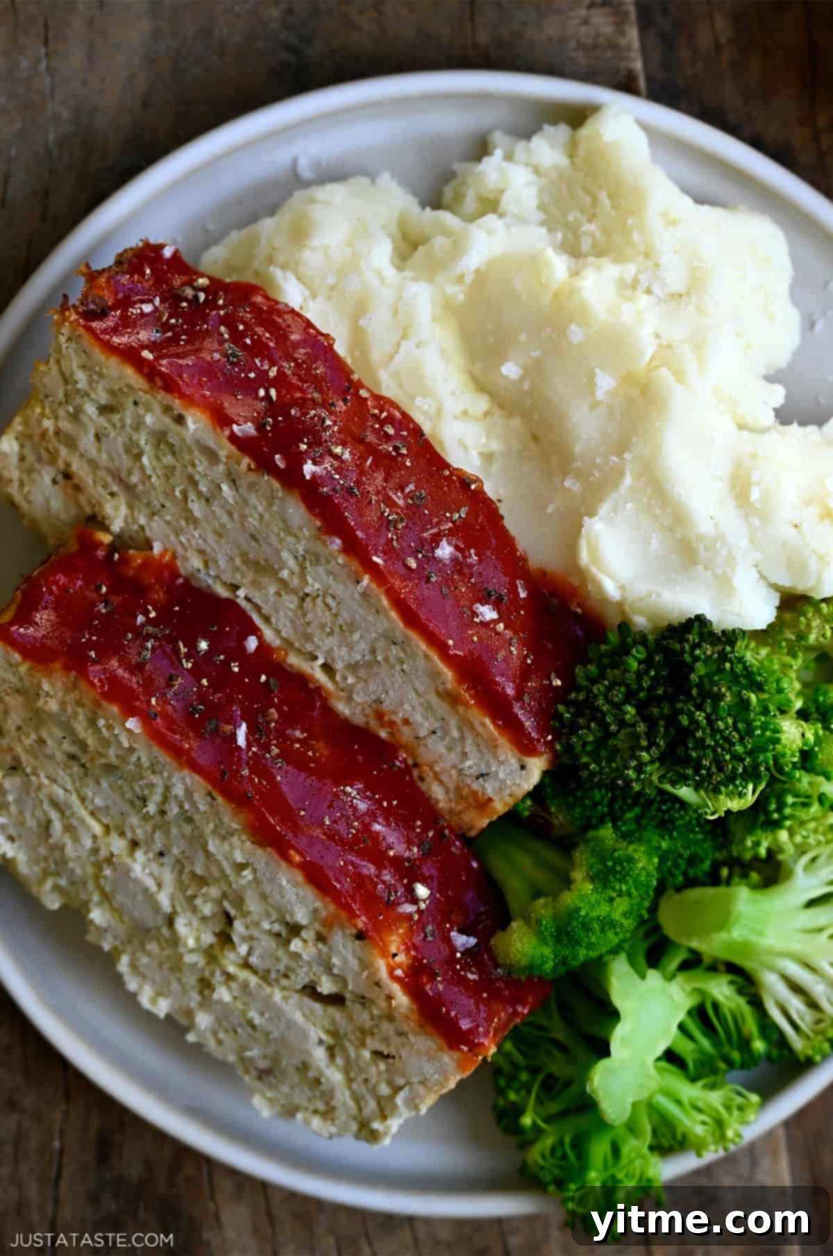 Two enticing slices of perfectly cooked ground chicken meatloaf, beautifully glazed with ketchup, resting on a plate alongside crisp steamed broccoli and creamy mashed potatoes. A complete and comforting meal.