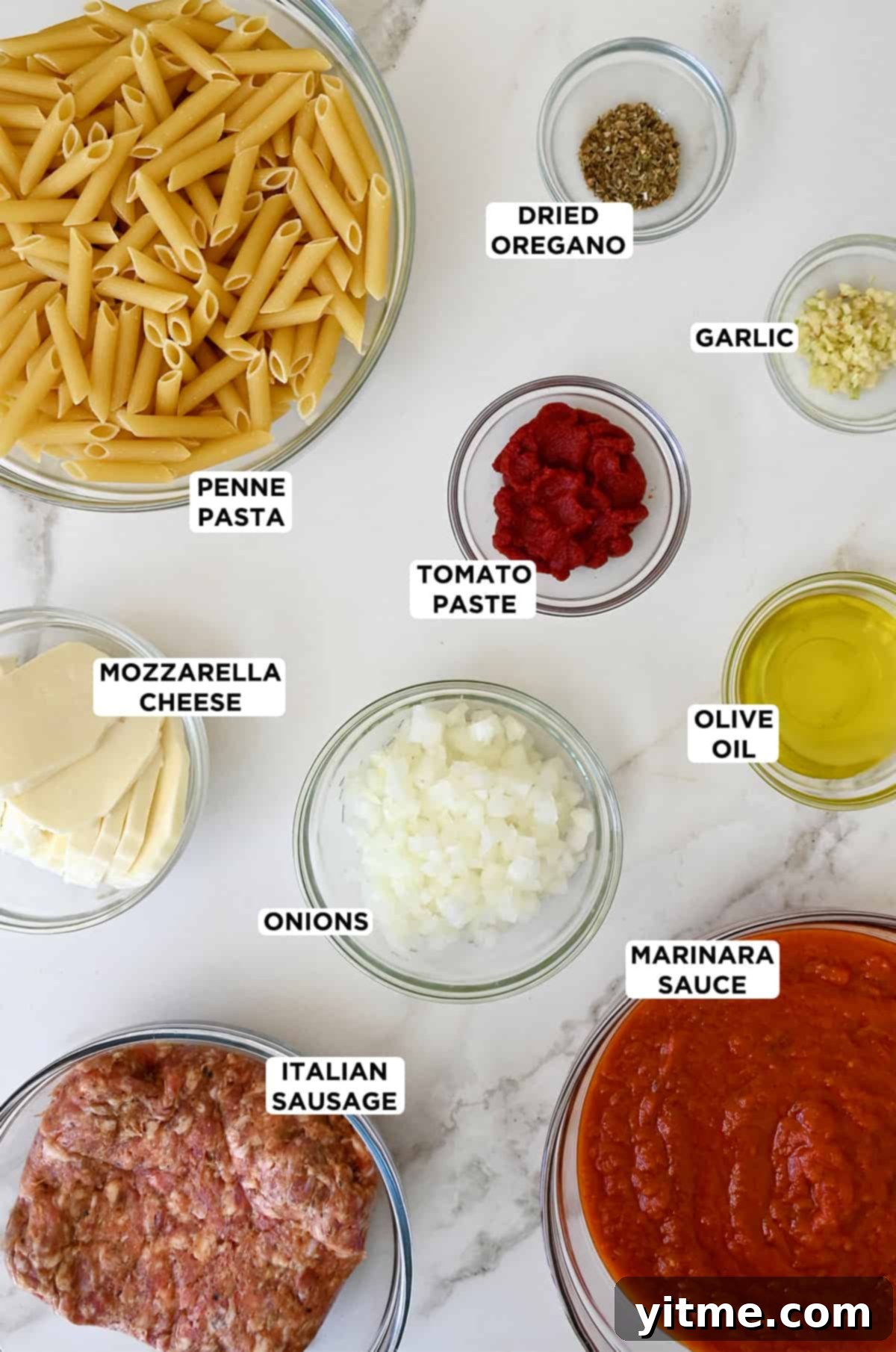 All the ingredients needed to make baked penne in clear glass bowls, including uncooked penne, dried oregano, minced garlic, tomato paste, olive oil, diced onions, marinara sauce, Italian sausage and slices of fresh mozzarella cheese.