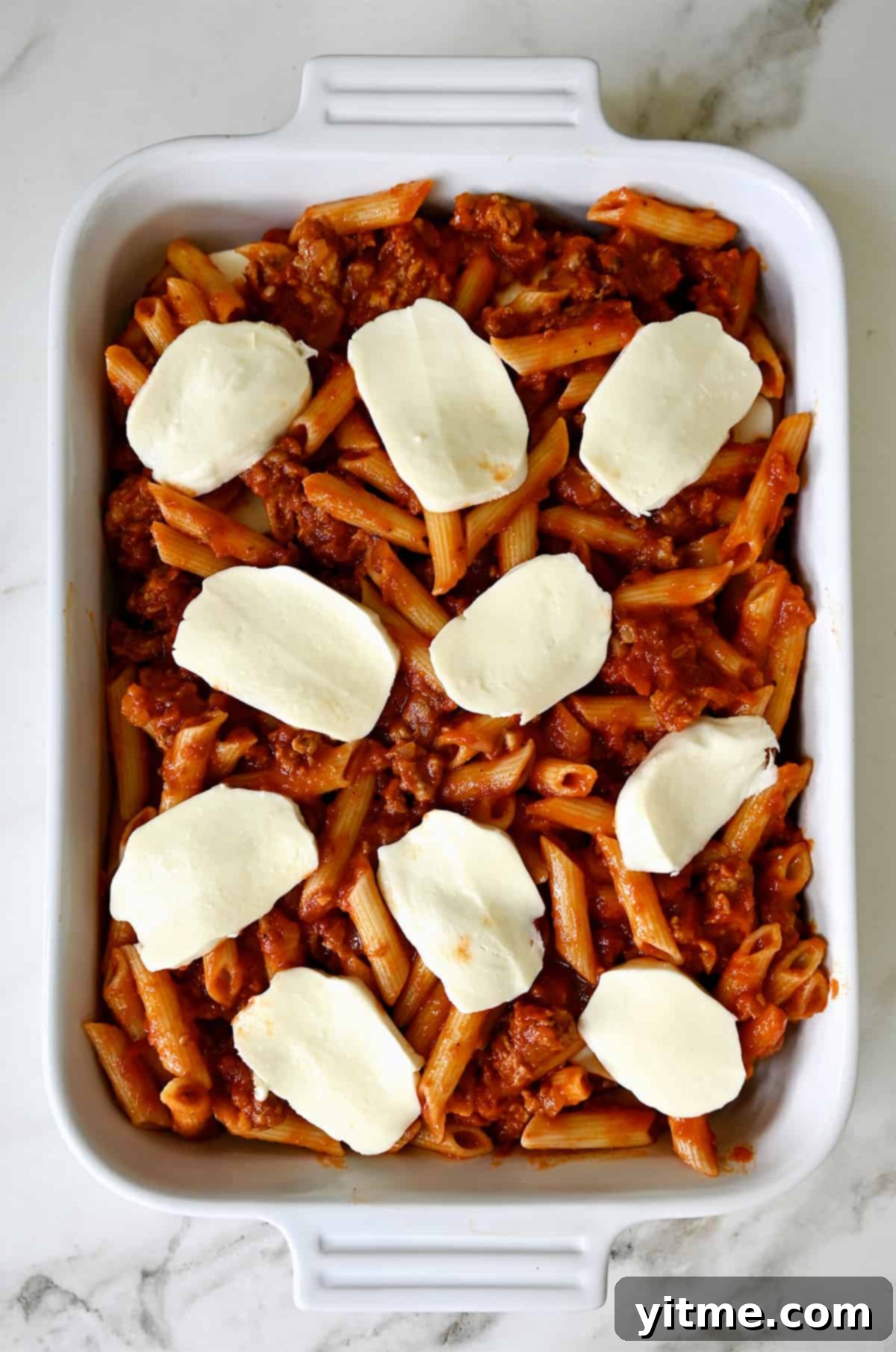 Slices of fresh mozzarella atop penne pasta with a bolognese sauce in a white baking dish.