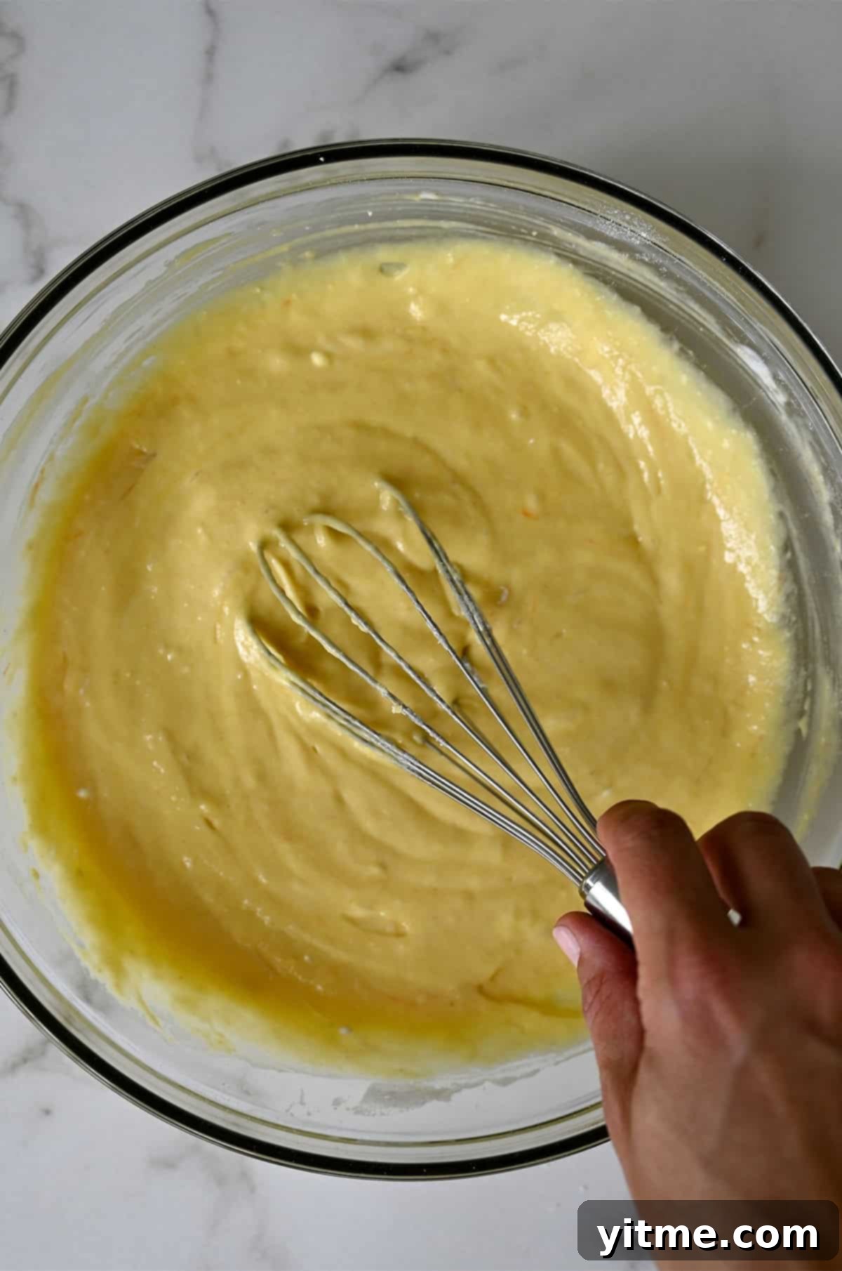 A whisk stirring vanilla cupcake batter in a bowl.