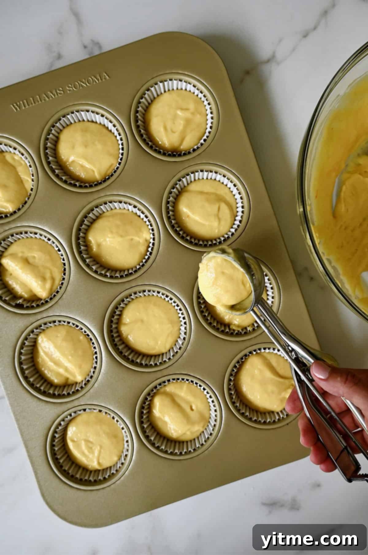 A hand holding a large cookie scoop adds cupcake batter to a muffin tin with liners.