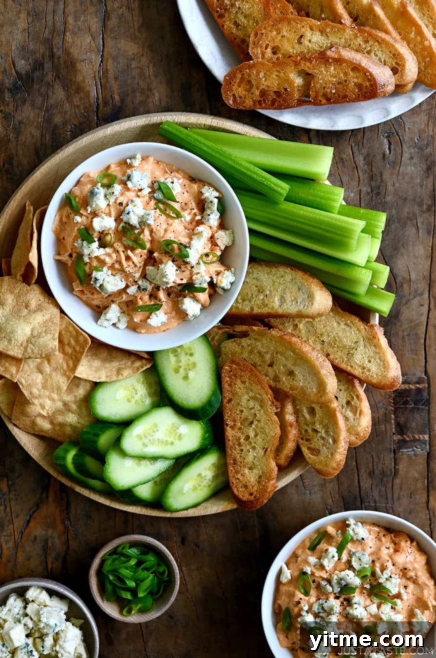 A top-down view of Slow Cooker Buffalo Chicken Dip in a small bowl on a plate surrounded by celery sticks, baguette toasts, sliced cucumber and tortilla chips