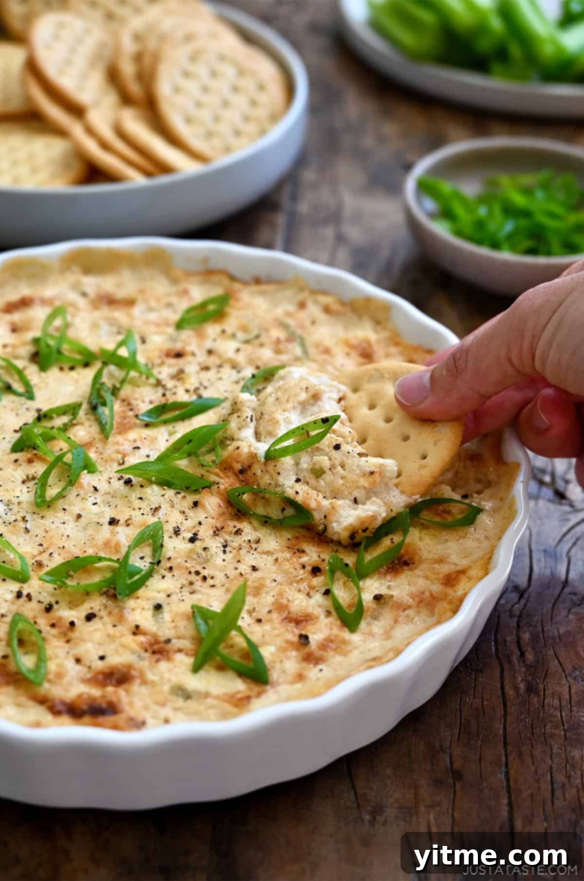 A hand dips a cracker into hot crab dip in a shallow serving dish.
