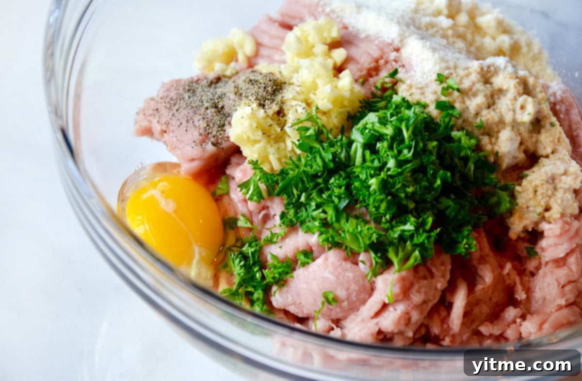 The ingredients for turkey meatballs are piled in a glass bowl, including ground turkey, egg, fresh parsley, breadcrumbs, minced garlic and seasonings.