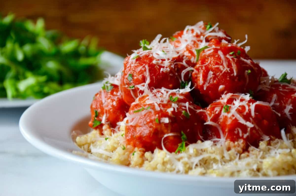 Baked turkey meatballs are coated in tomato sauce and sprinkled with parmesan cheese. They sit on a bed of quinoa in a white bowl. A white dish of greens is in the background.