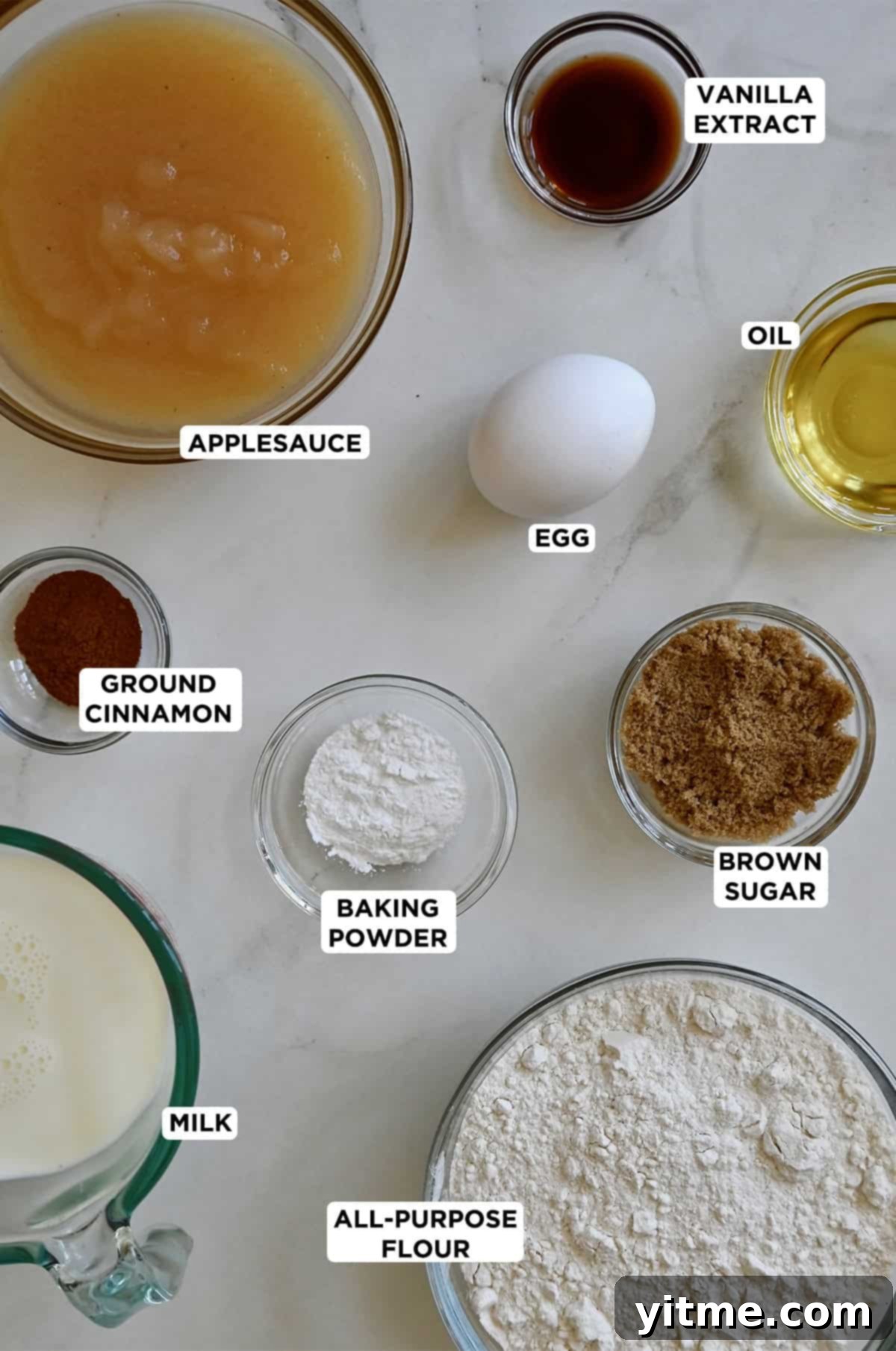 Various sizes of glass bowls containing the ingredients to make apple pancakes, including applesauce, flour, ground cinnamon, brown sugar, an egg, oil, baking powder and milk.