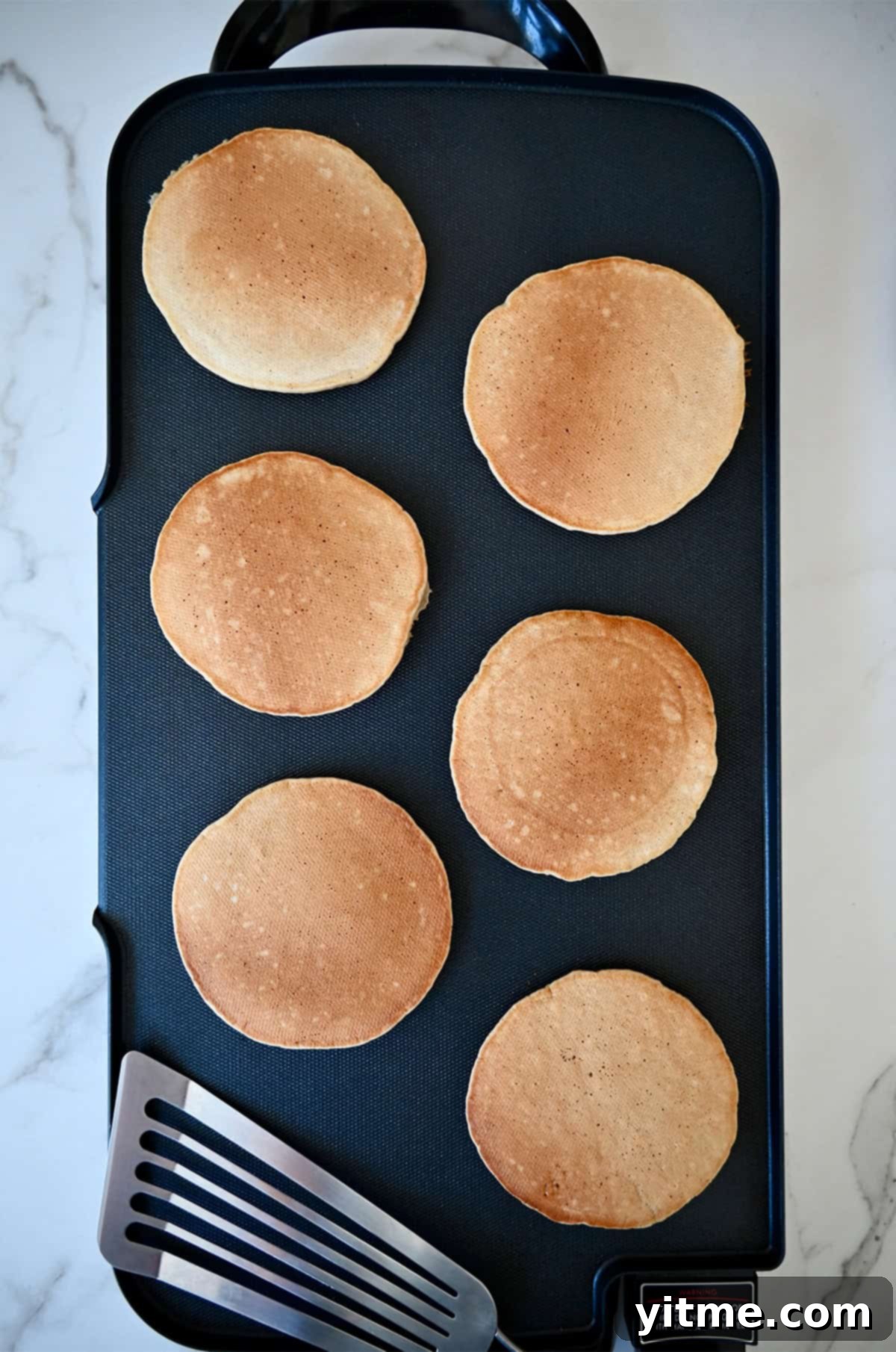 Golden brown apple cinnamon pancakes on a griddle next to a fish spatula.