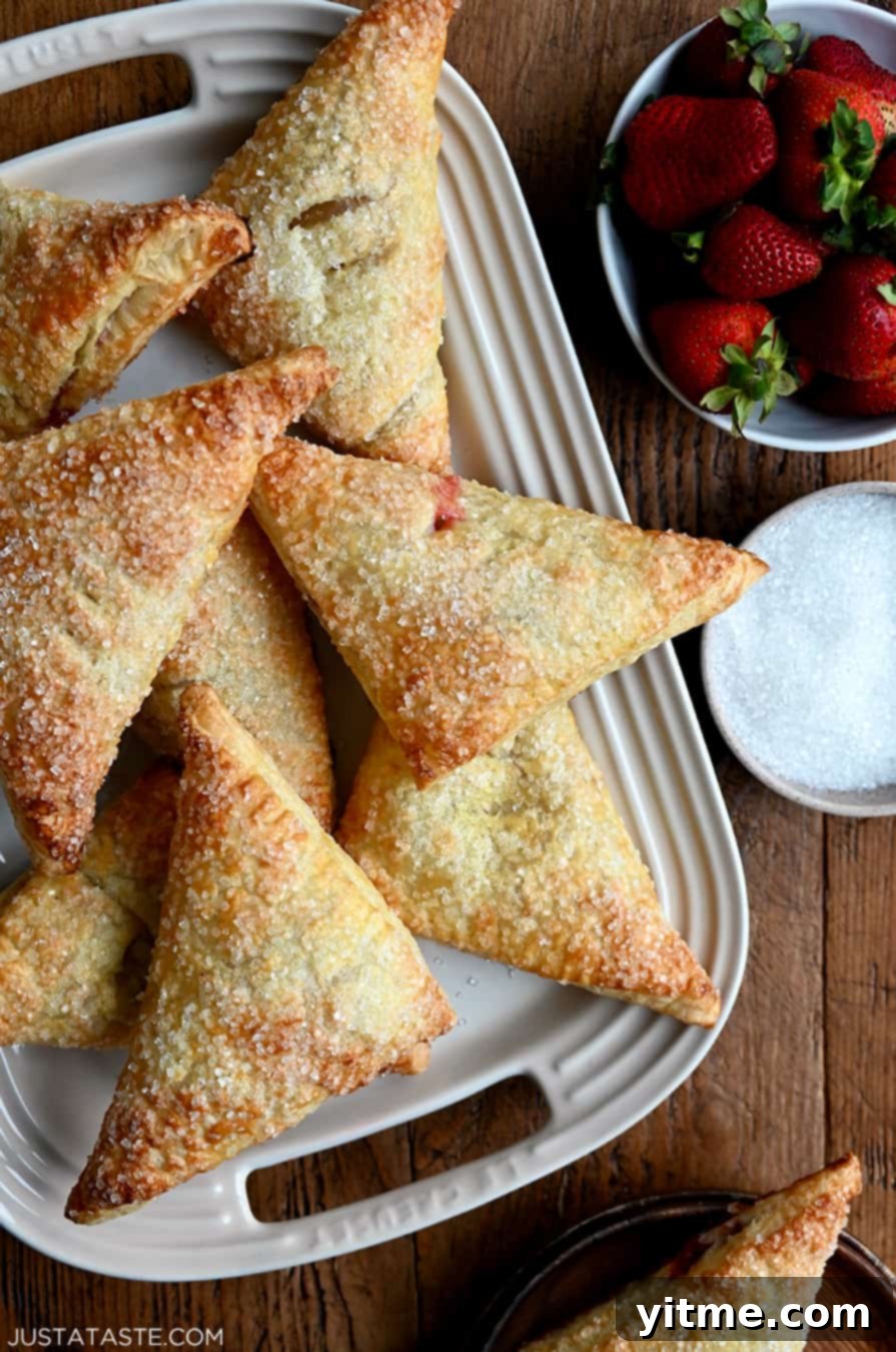 Strawberry Rhubarb Turnovers on a white serving plate next to a bowl containing fresh strawberries and a bowl containing sanding sugar