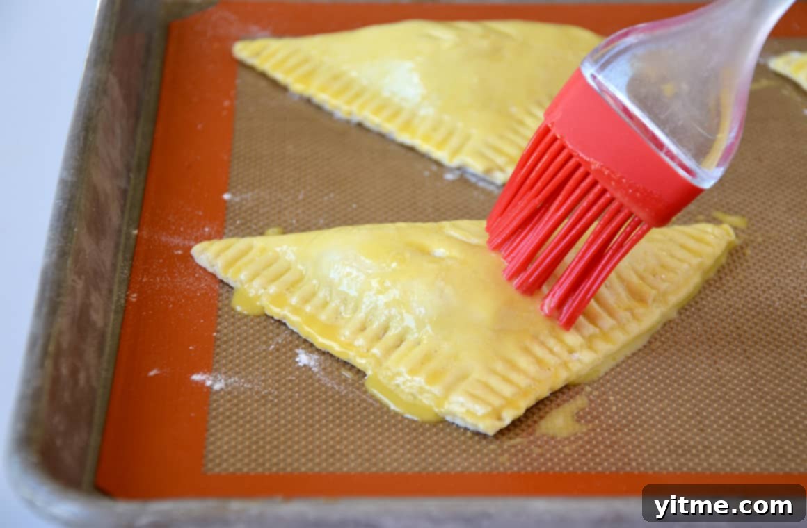 A pastry brush applies an egg wash atop unbaked pastries on a lined baking sheet