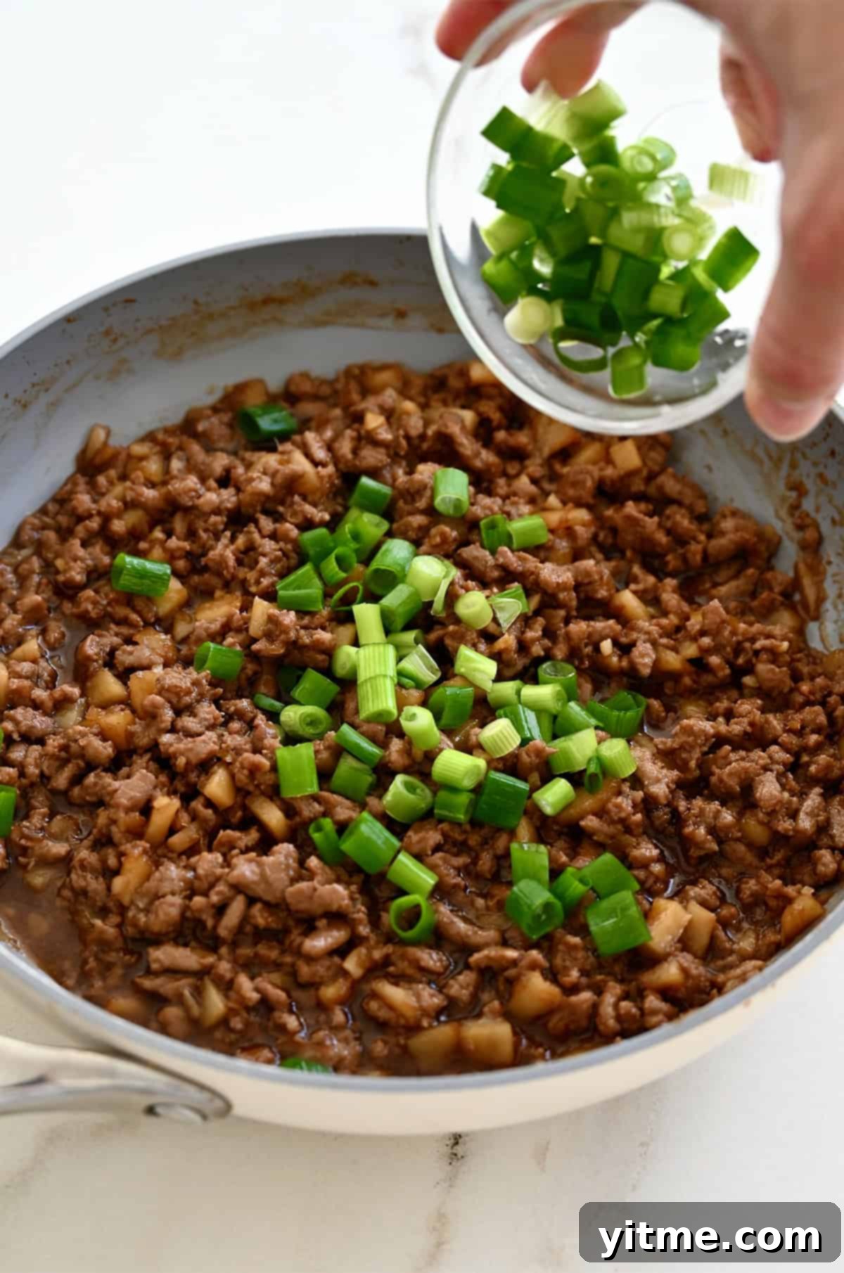 Chopped scallions from a small bowl being added to a skillet containing Chinese chicken.